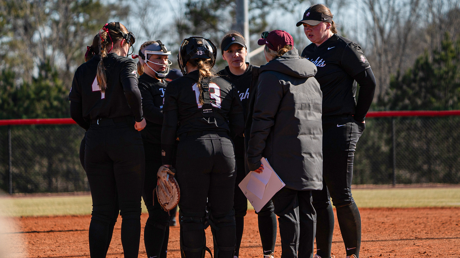 Softball team huddles on the field with head coach Whitney Arion