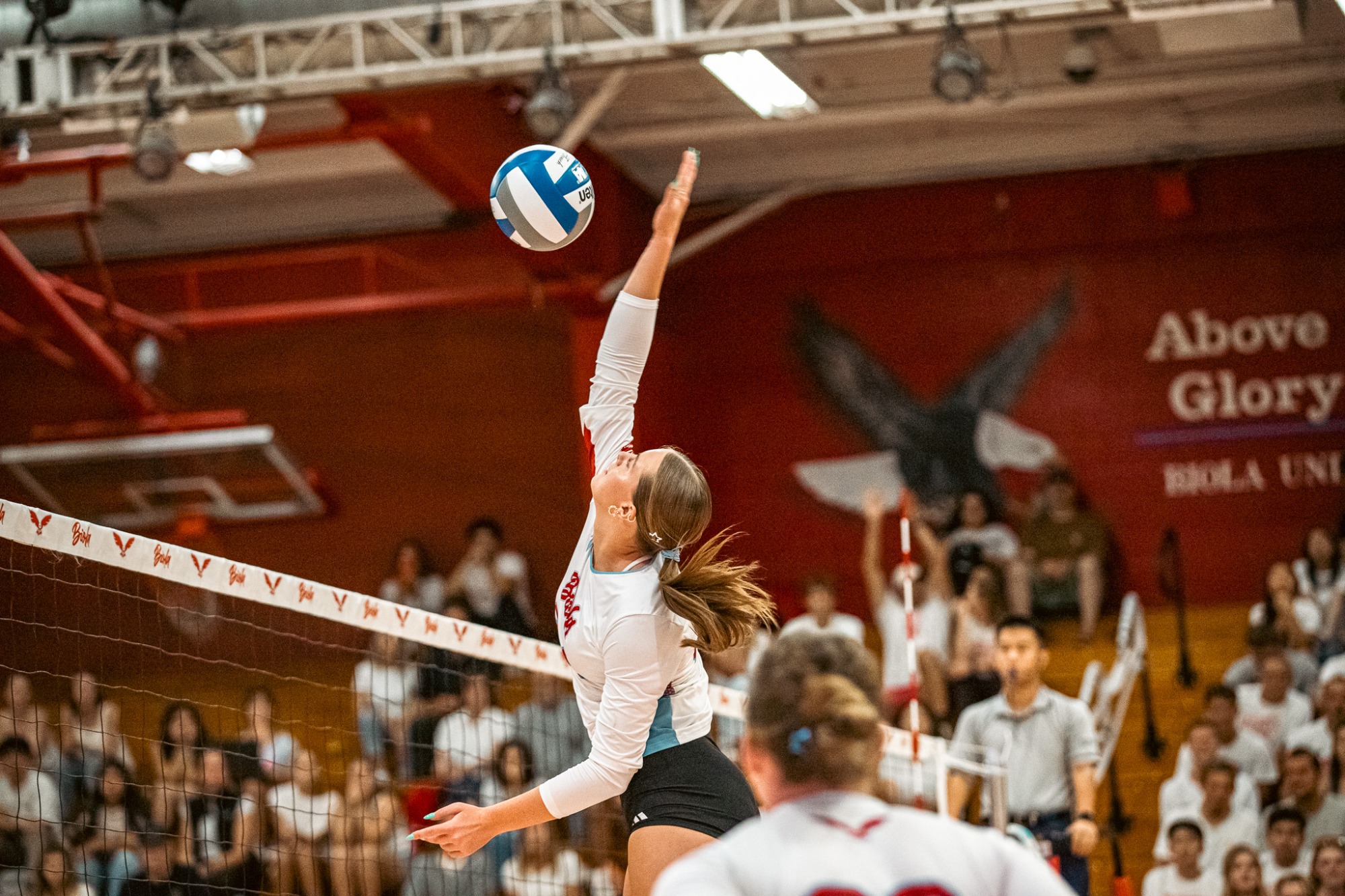 A Biola hitter rises above the net and reaches to attack as the ball hangs over her hand.