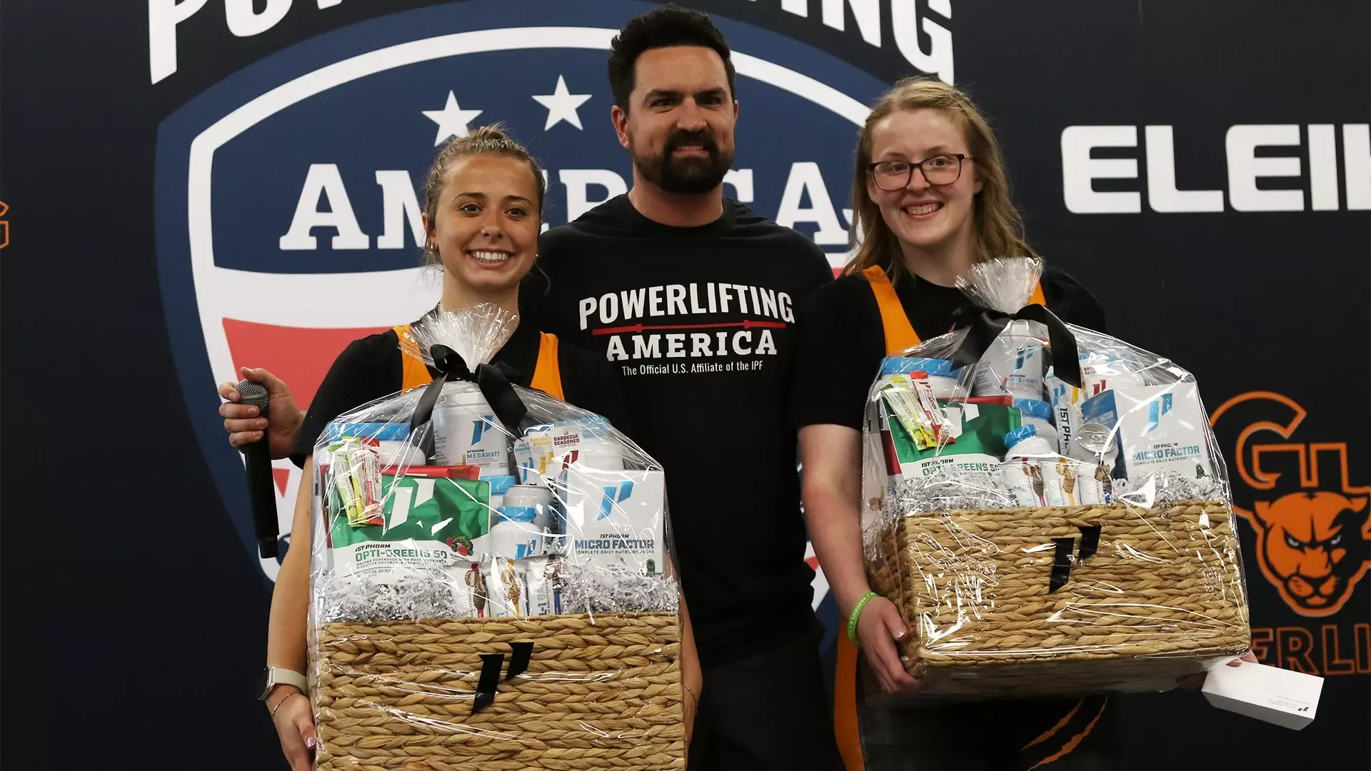 Three people stand together. Two girls with blond hair, one with glasses, hold gift baskets. A man with dark hair and a beard stands between them.