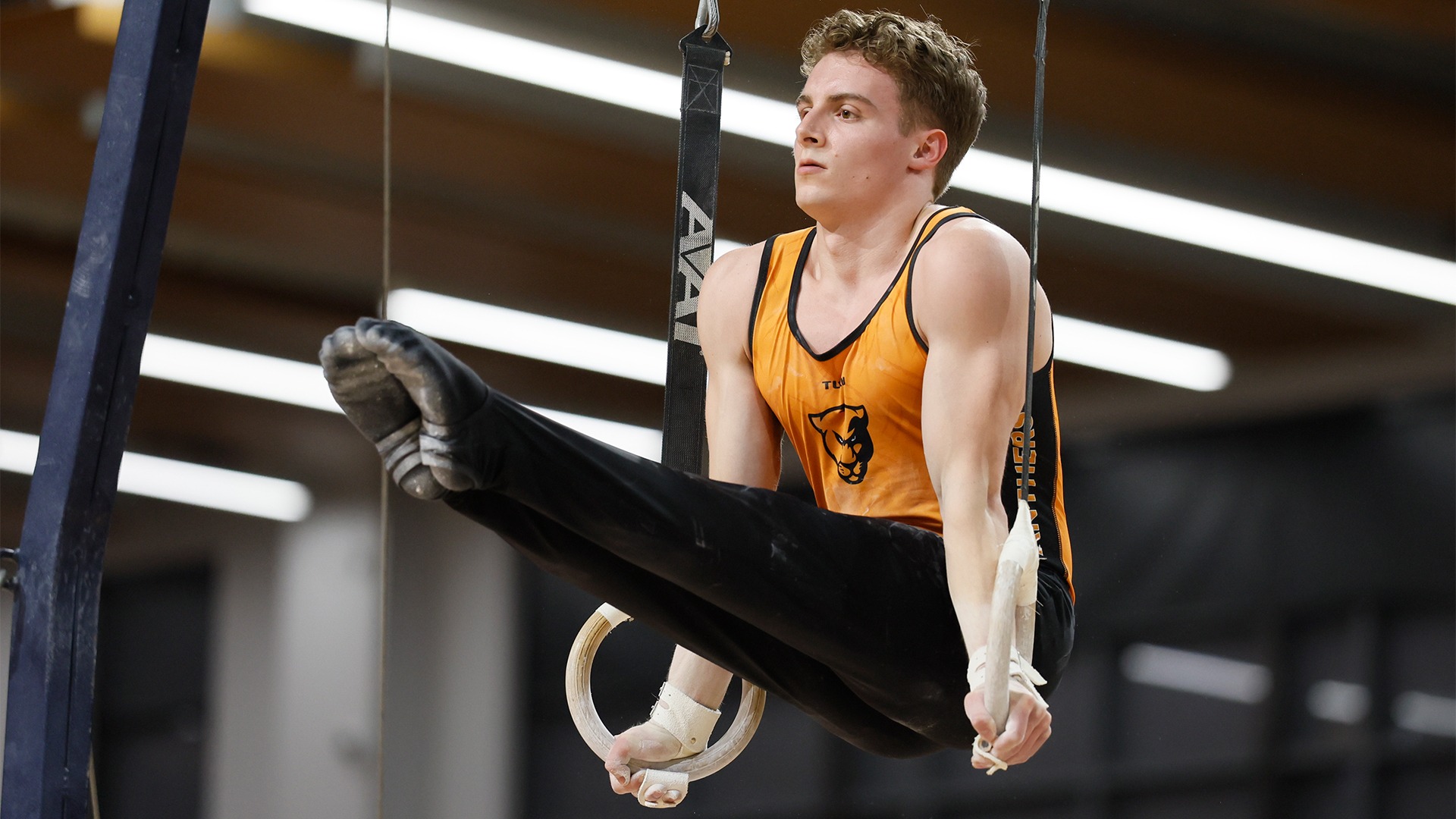 Ethan Poole in orange jersey, black pants on rings with feet straight out