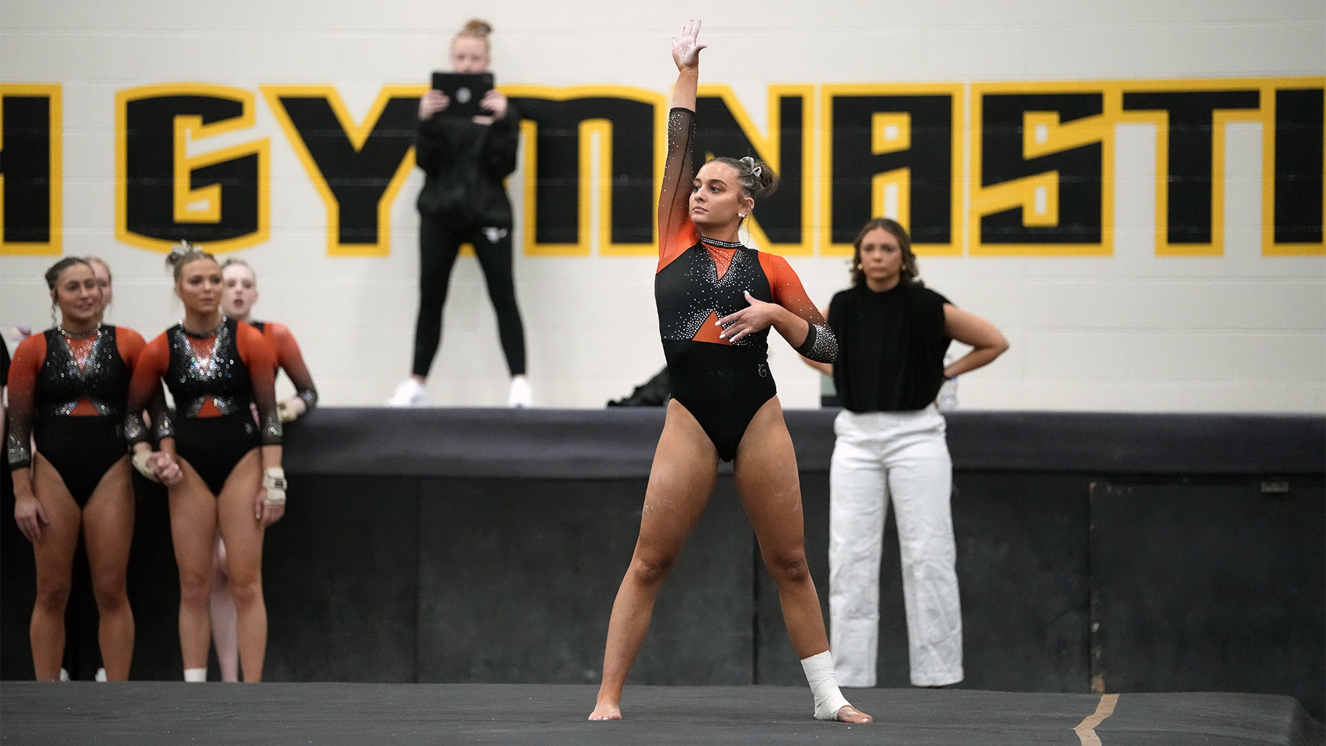 Women's gymnast in black and orange leotard on floor with hand in air