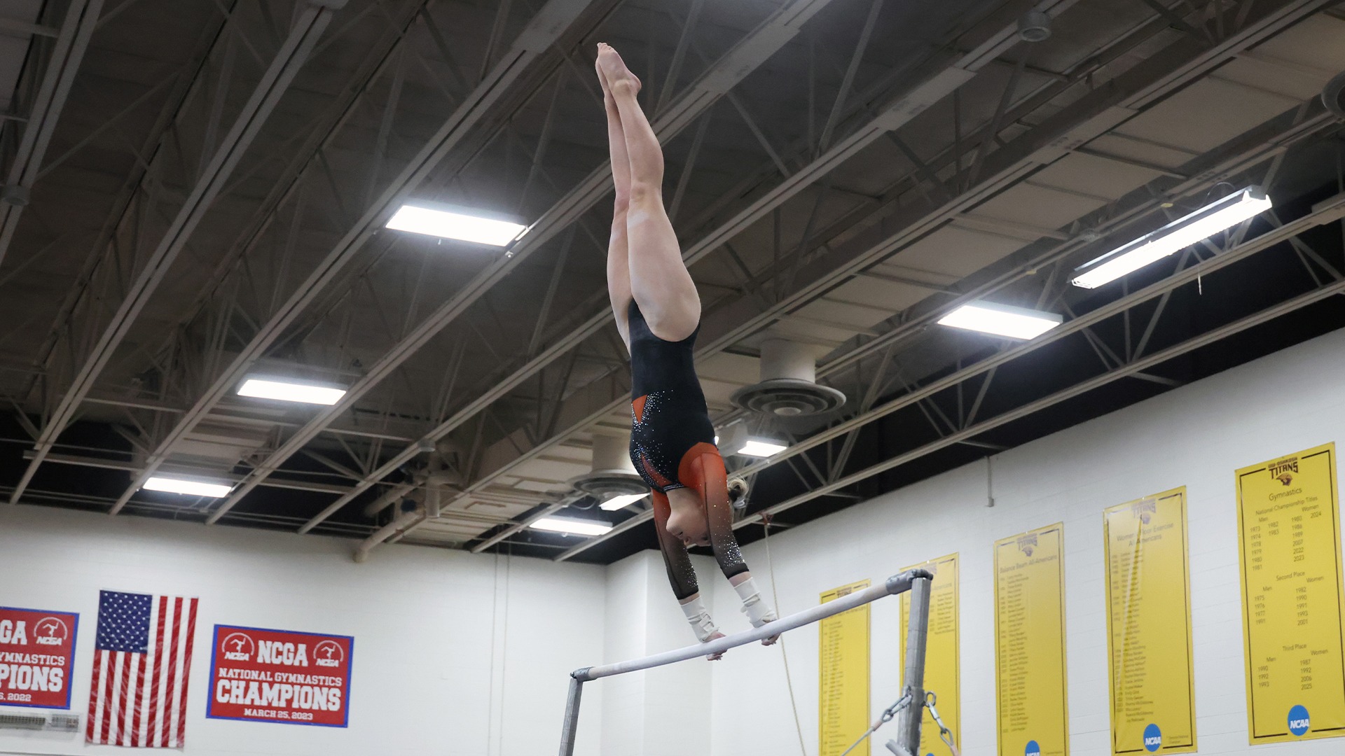GU Woman gymnast in orange and black leo upside down on bars