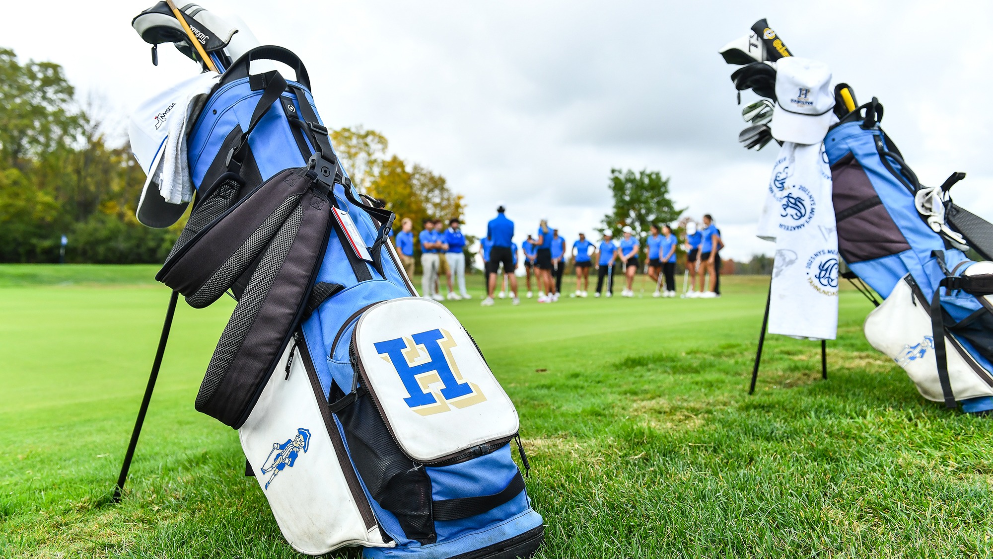 Two Golf bags propped up near the practice green in the foreground, Coach Lauren Cupp talks with the teams in the background