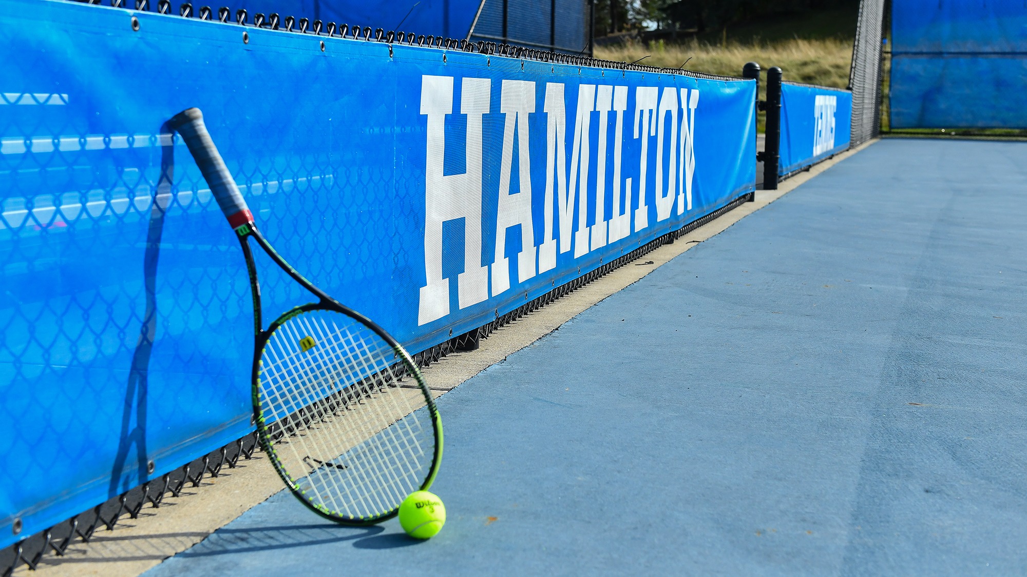 Tennis racket leans against the fence in front of the blue Hamilton banner