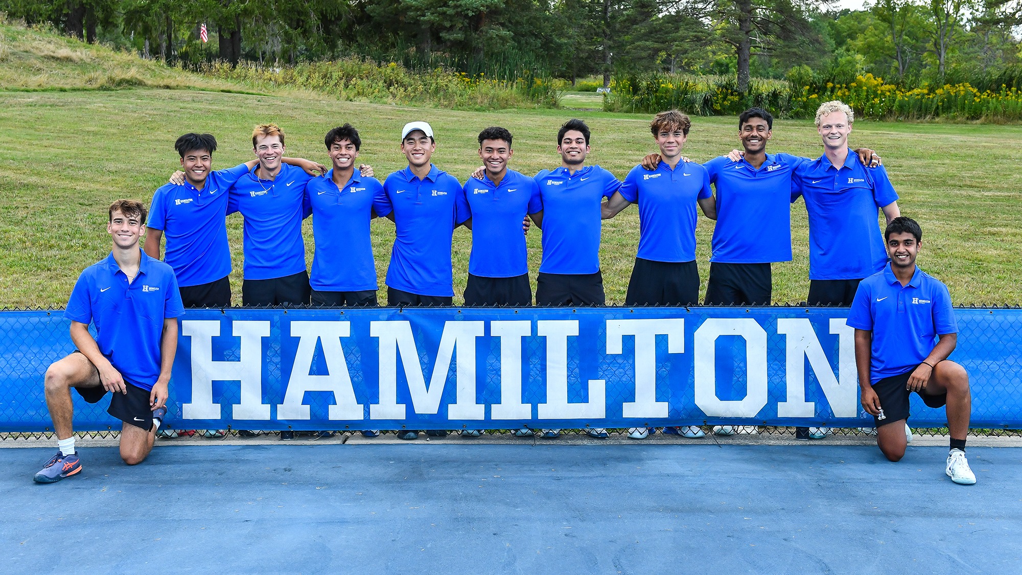 Men's Tennis players pose for a team photo around a Hamilton banner at Tietje Family Tennis Center