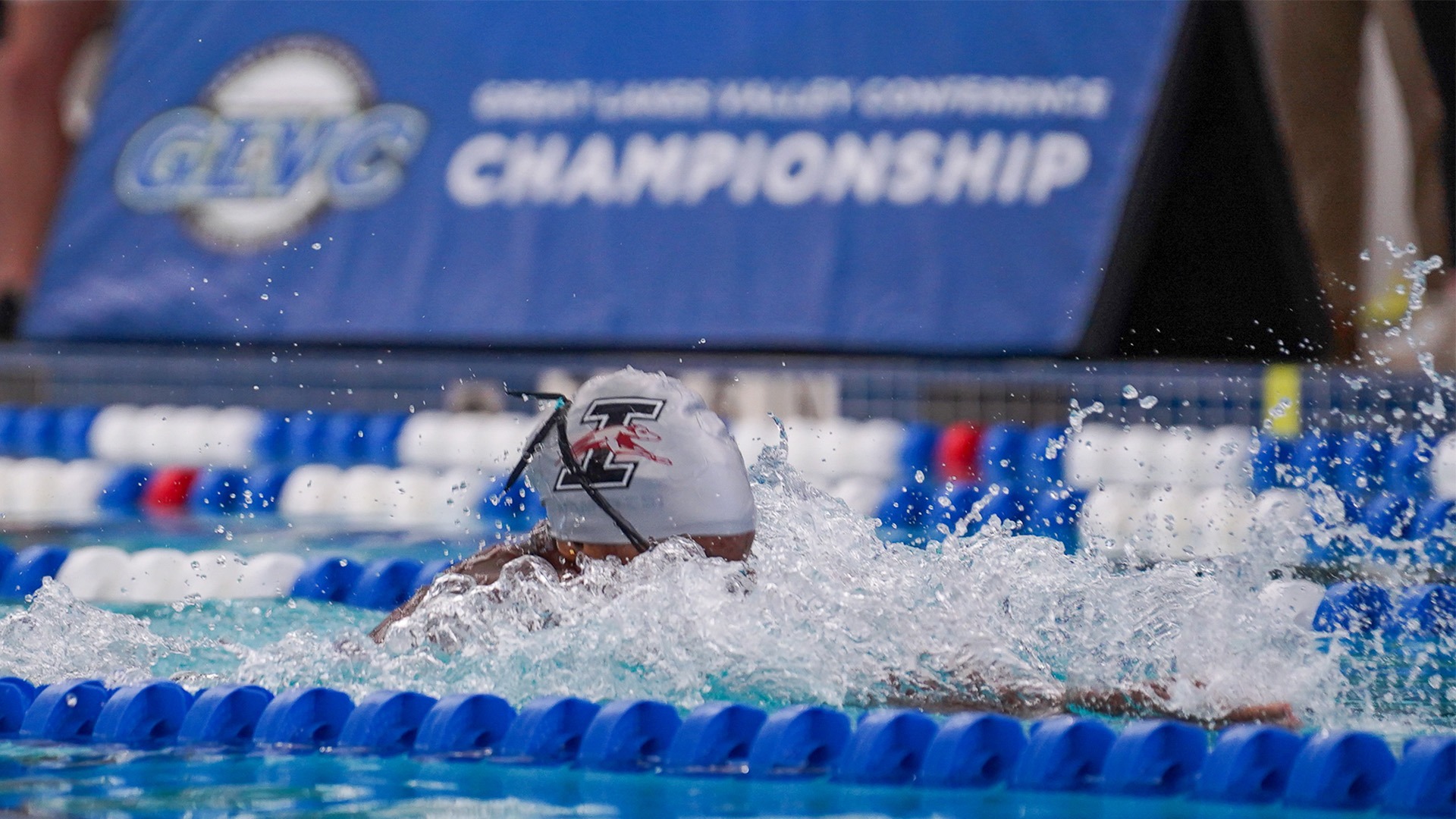 A UIndy swimming student-athlete is shown competing with only their UIndy swim cap visible. The GLVC Championships logo is shown out of focus in the background.