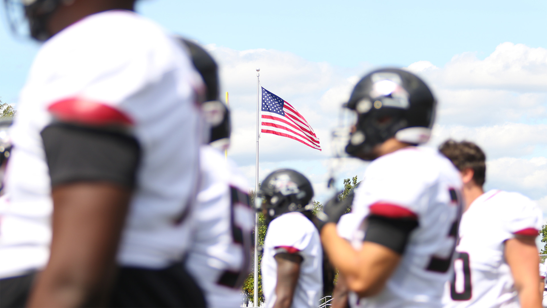The America flag waves in the background as UIndy football players watch a game from the sidelines.