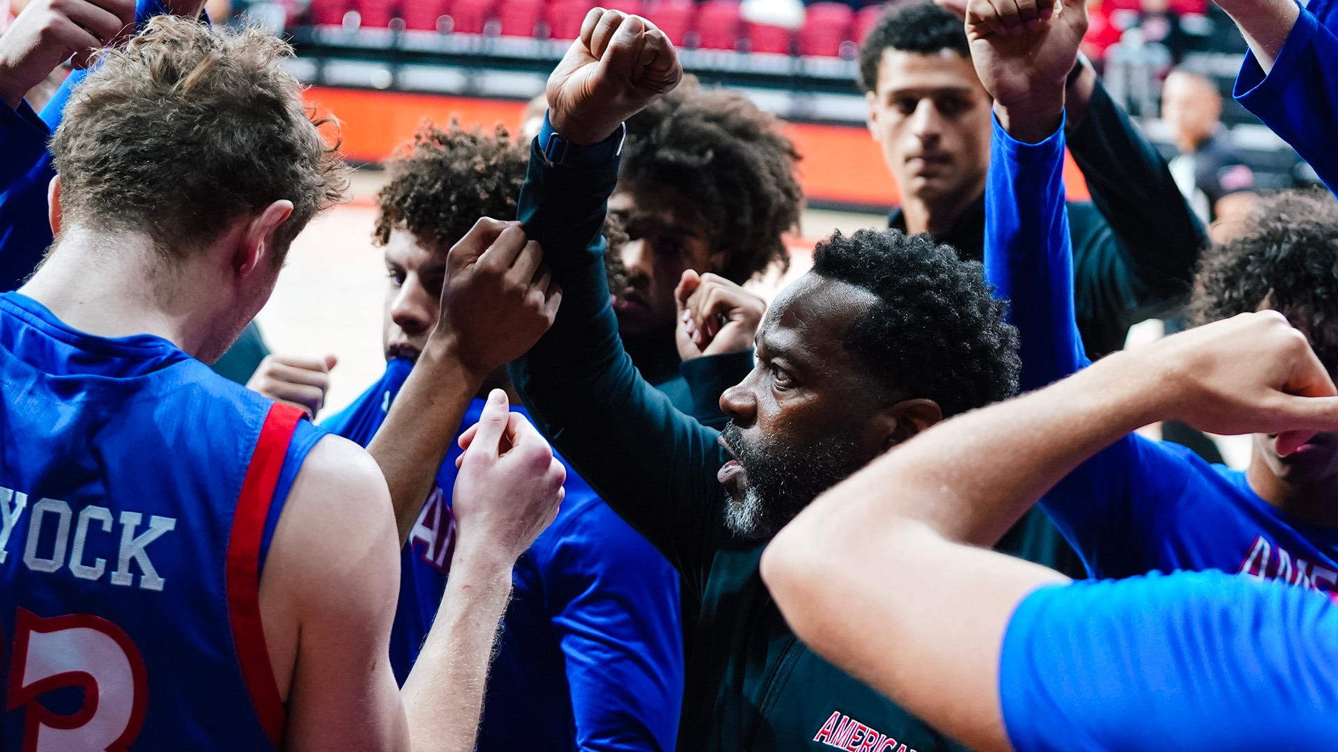 Coach Duane Simpkins in the middle of a hands up huddle with men's basketball