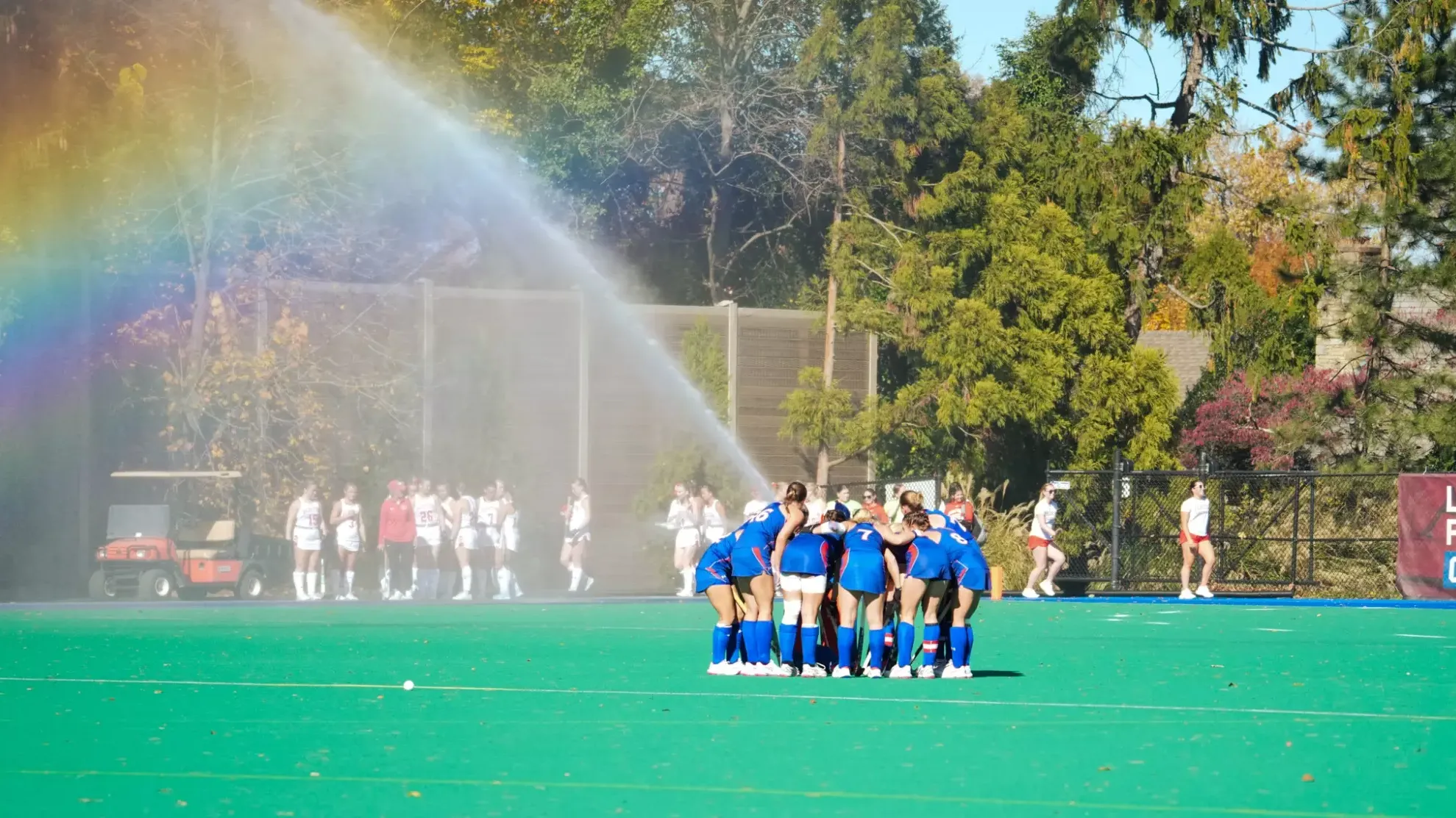 The field hockey team huddled together pregame with the water cannon creating a rainbow in the background.