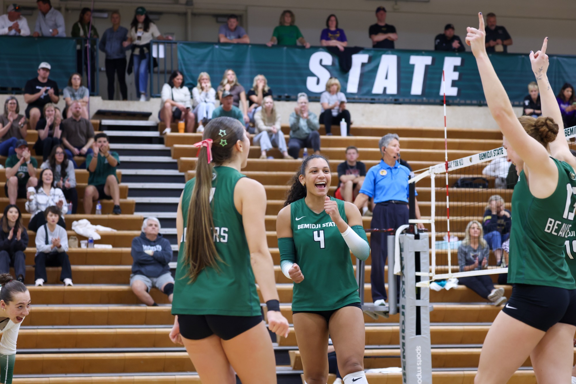 Bemidji State volleyball players celebrate near the net as spectators applaud from the bleachers.