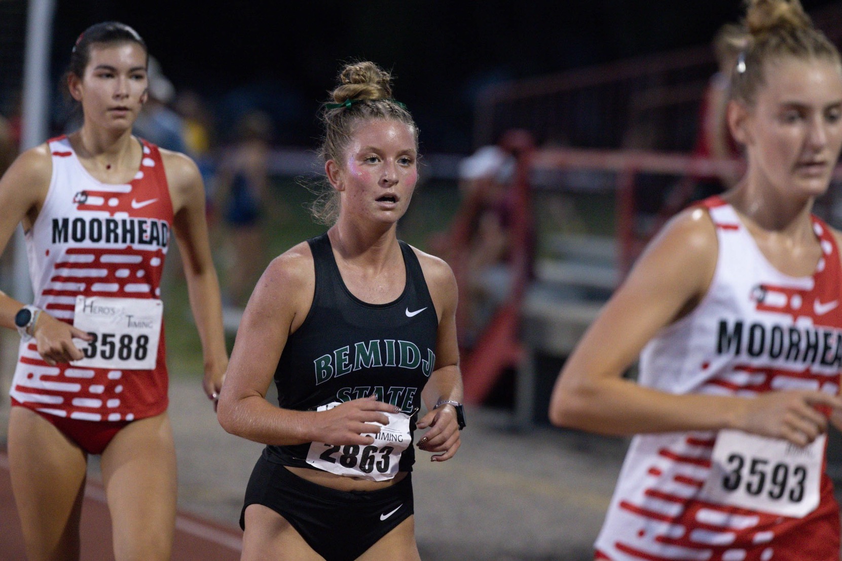 A BSU runner races in a black tank top with green and white letters. Her hair is pulled back into a bun and she runs between two competitors. 