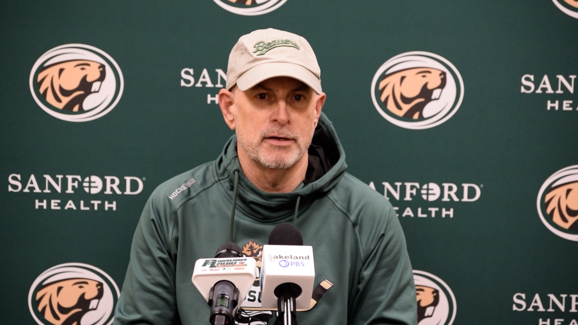Tom Serratore wearing a green Bemidji State Beavers hoodie and a beige Beavers baseball cap sits at a press conference table. Multiple microphones with station logos, including Lakeland PBS, are positioned in front of them. The backdrop features repeated Bemidji State Beavers logos and “Sanford Health” branding.