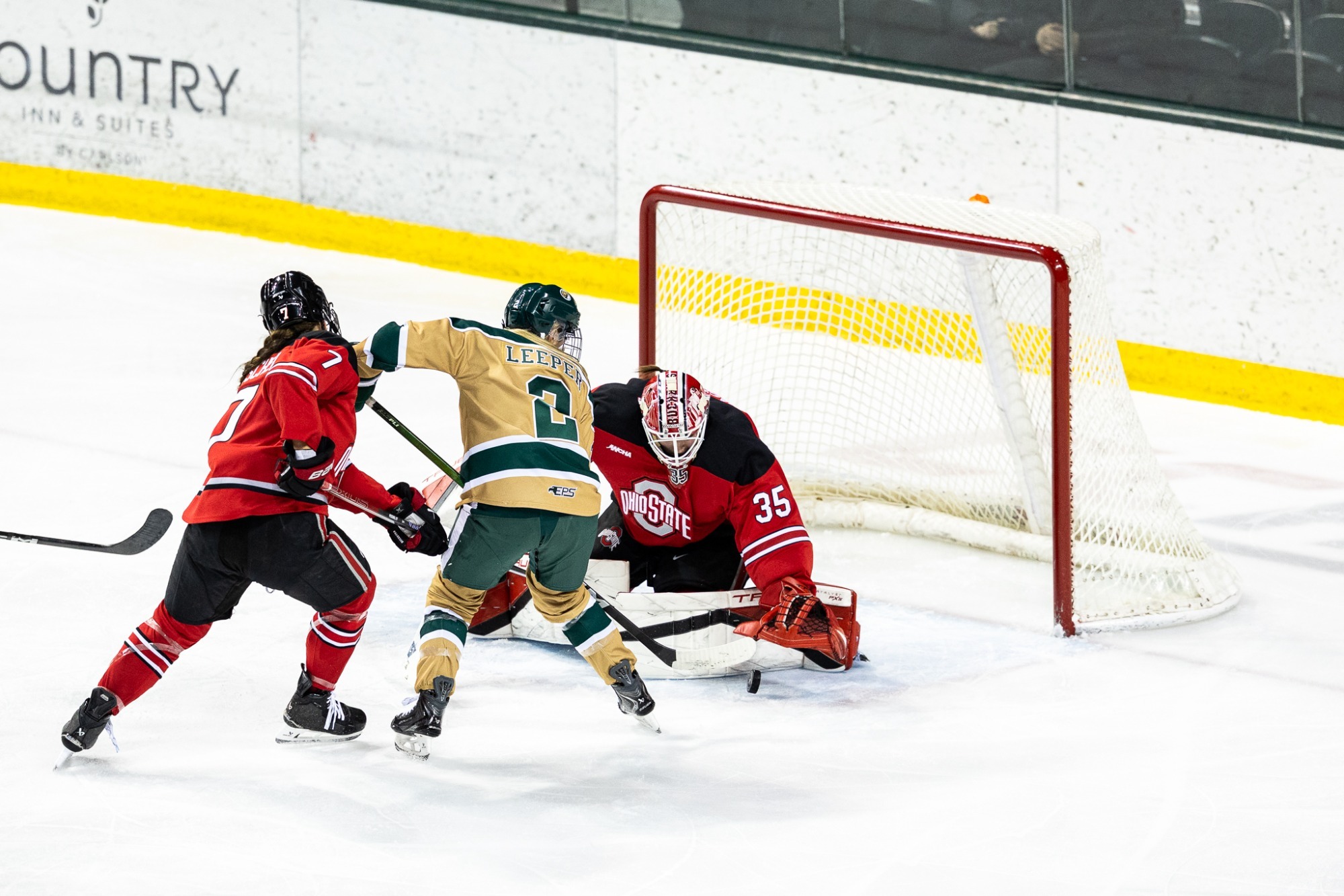 A hockey player in tan and green takes a close shot on goal while an Ohio State goalie in red and black crouches to block it, with a defender beside the shooter. The action occurs directly in front of the net.
