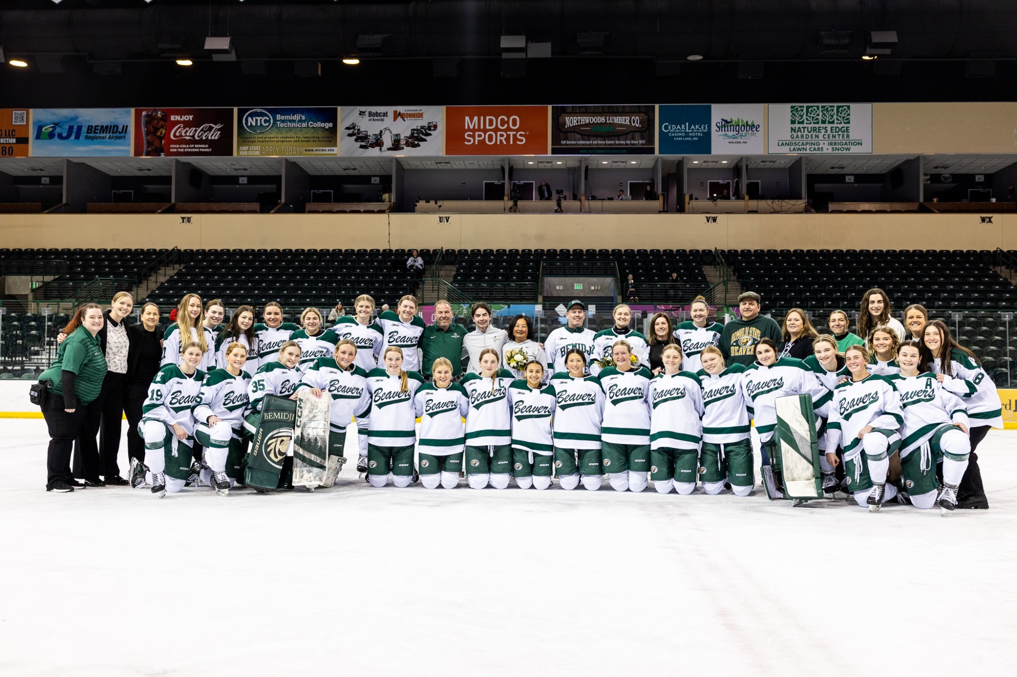 A large group photo of a women’s ice hockey team and staff posing together on the ice inside an arena.