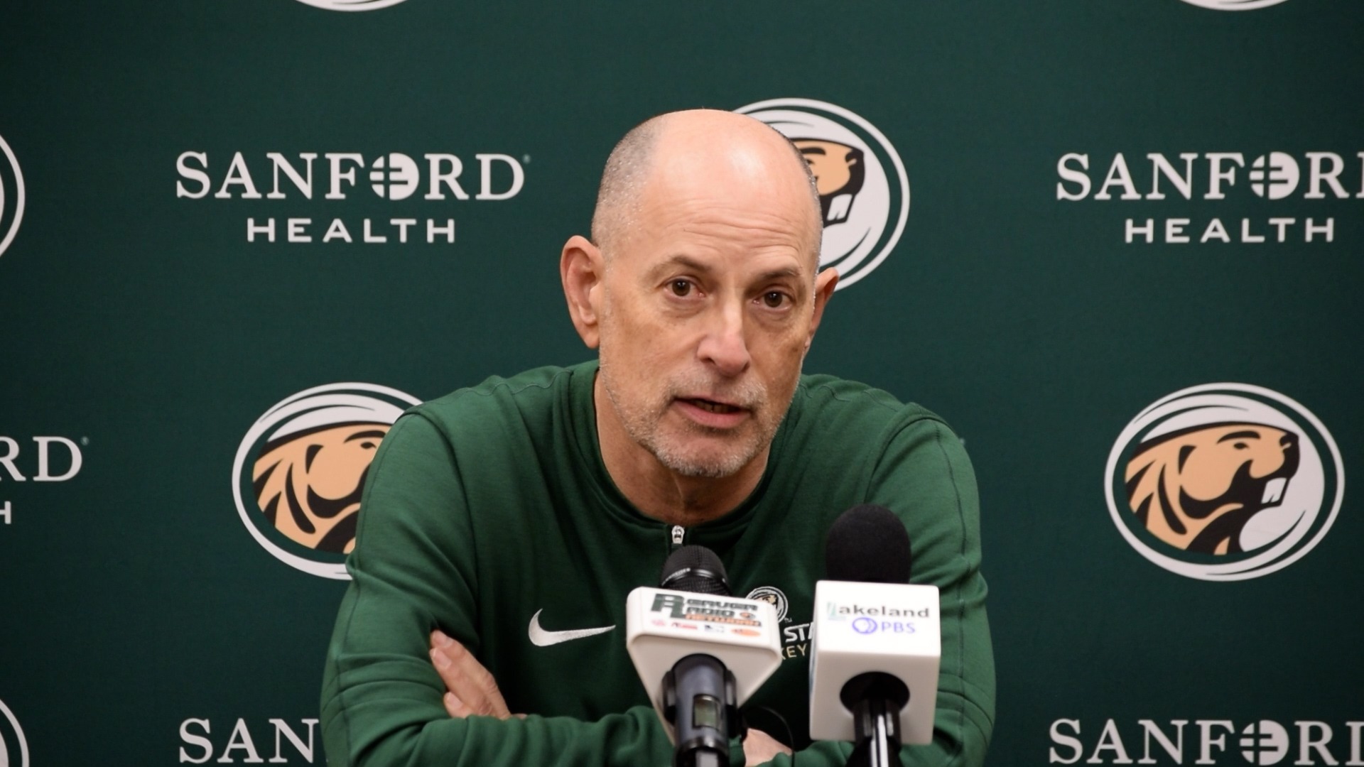 Tom Serratore in a green athletic pullover is seated at a press conference table with multiple microphones in front of them. The backdrop features Bemidji State University beaver logos and Sanford Health branding.