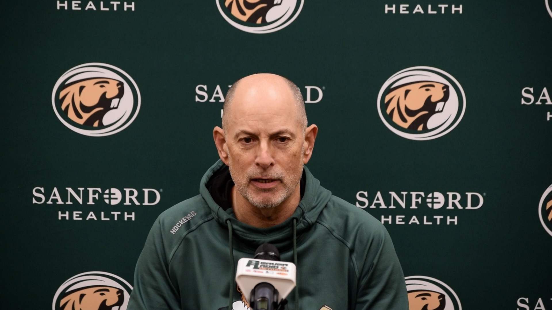 A person wearing a green athletic hoodie speaks at a press conference podium with a microphone. The backdrop features repeated Bemidji State–style beaver logos and Sanford Health branding.
