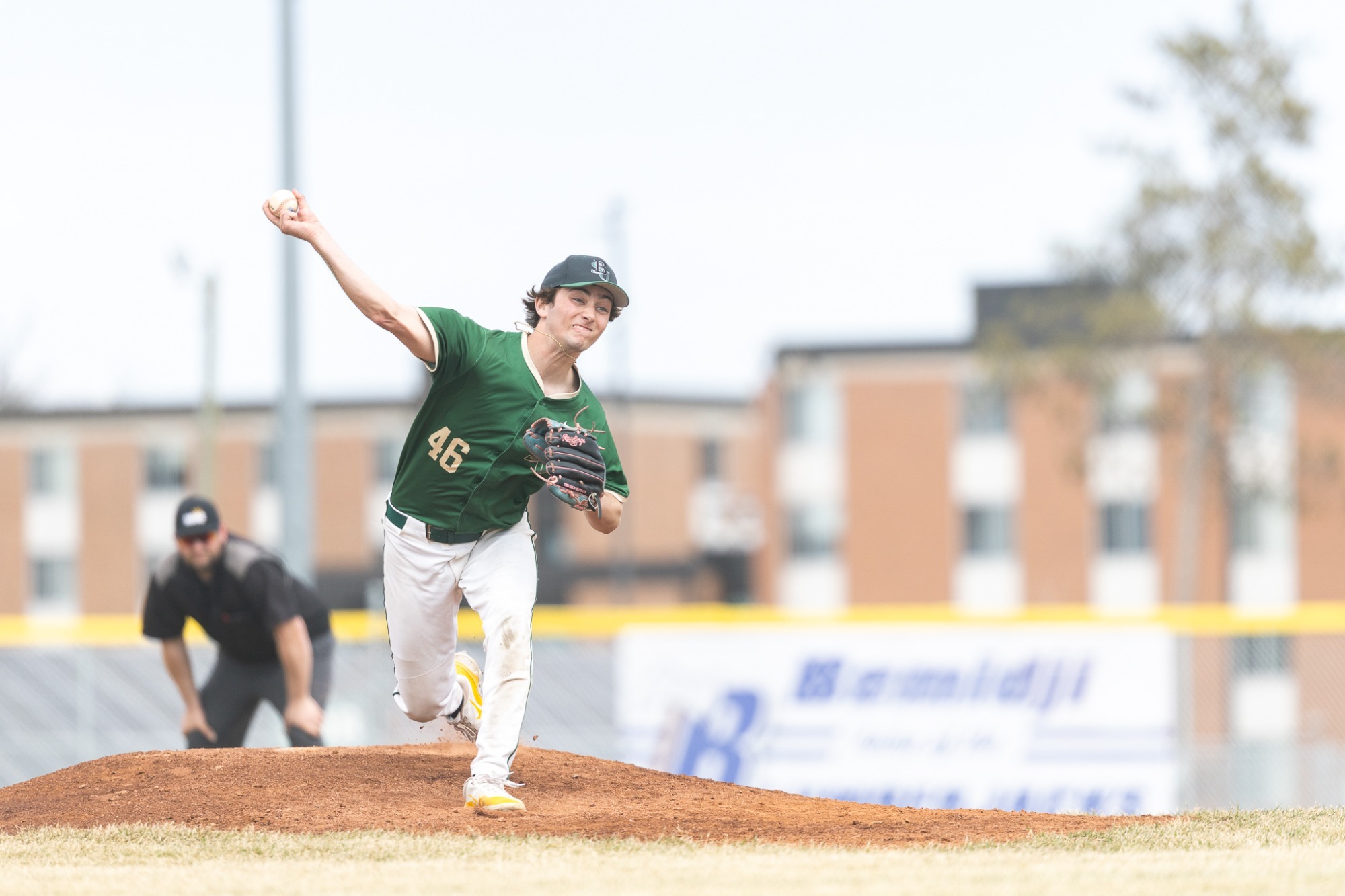 Andrew Murphy releases the baseball, leaning forward with the throwing arm fully extended and the back foot lifted off the mound. An umpire and parts of the field are visible behind the pitcher, slightly blurred.