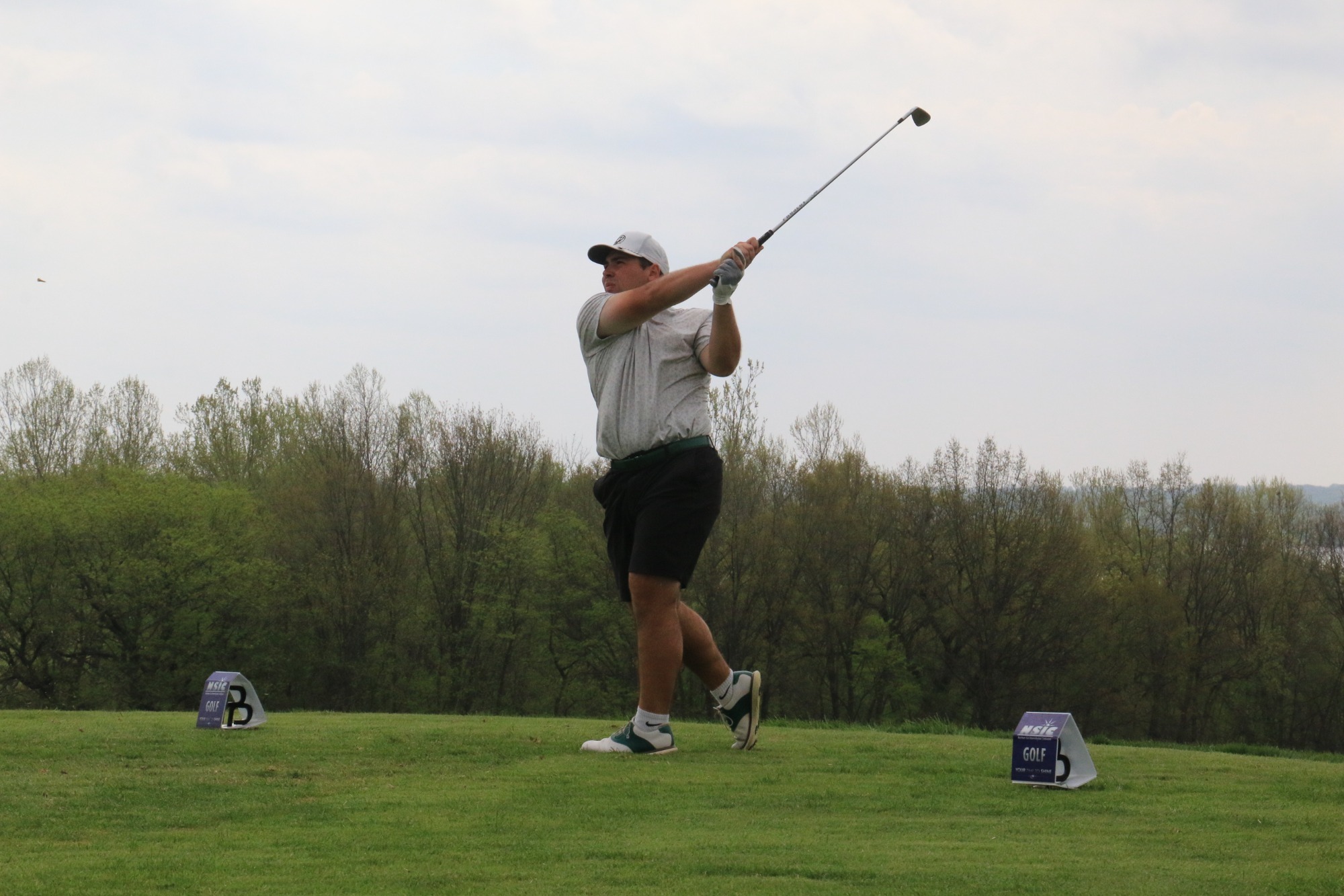 Logan Schoepp finishes a swing on a grassy tee box, the club raised over the shoulder. Tee markers sit nearby, with trees and an overcast sky in the background.