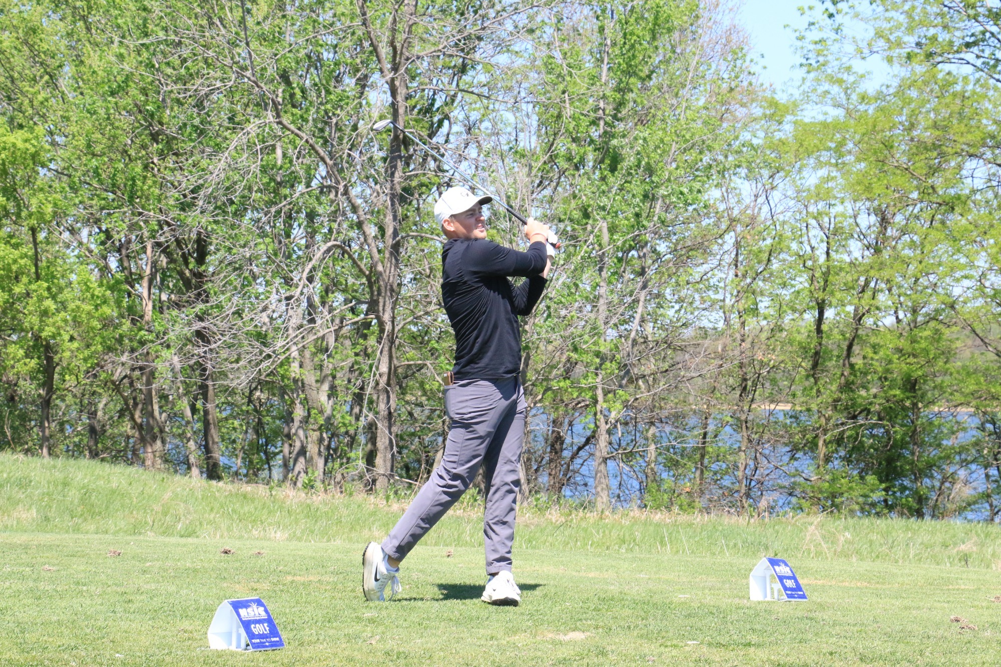 Teagan LaPlante completes a swing on a grassy tee box, the club raised overhead in follow-through. Tee markers sit on the grass, with trees and a body of water visible in the background under a bright sky.