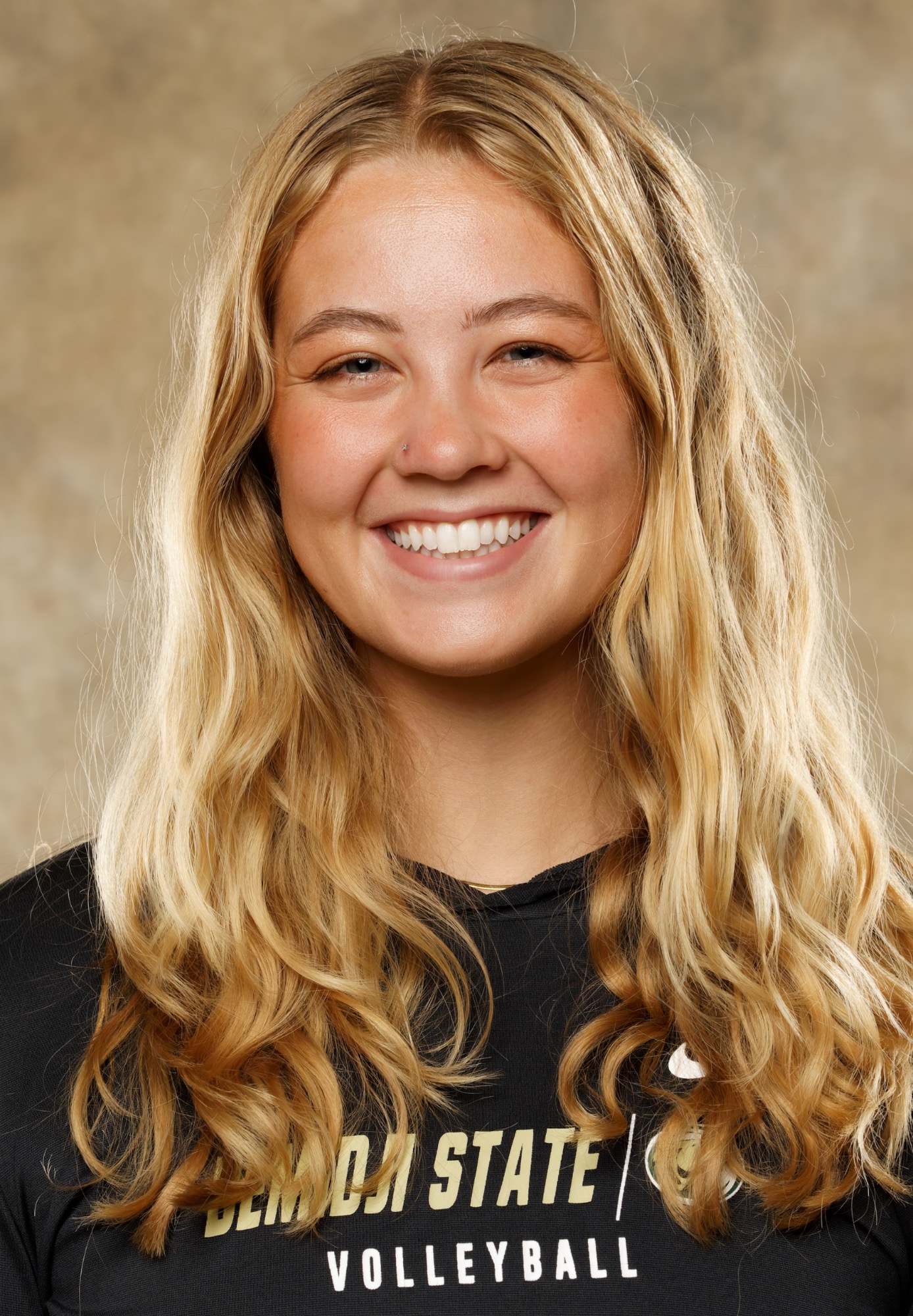Studio headshot of a Bemidji State volleyball coach wearing a black team hoodie. The coach has long wavy blonde hair and a small nose stud, photographed against a neutral studio backdrop.