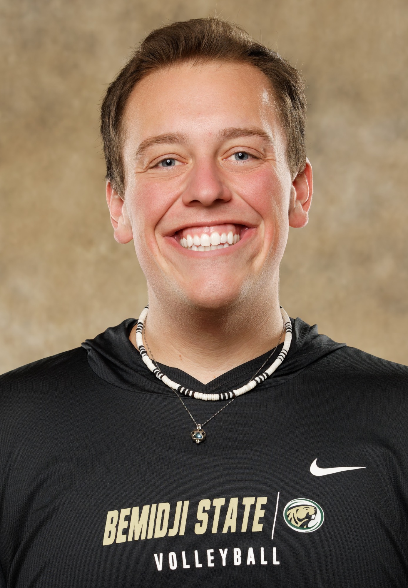 Studio headshot of a Bemidji State volleyball coach wearing a black team hoodie. The coach has short dark hair and layered necklaces, posed against a neutral studio background.