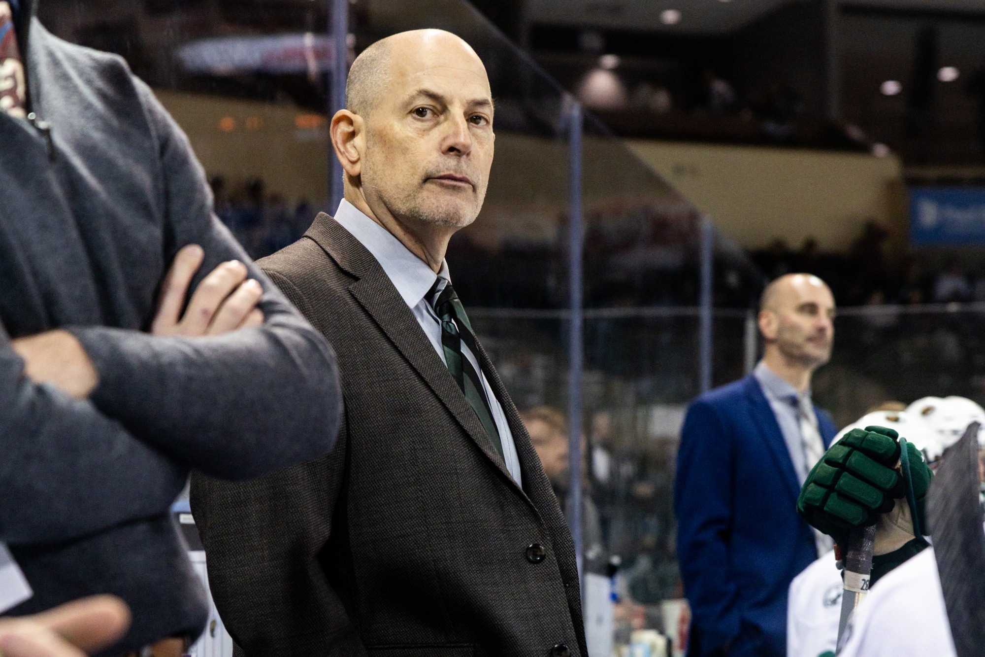 Tom Serratore stands behind the bench during a game, watching the action on the ice while assistant coaches and players stand nearby.