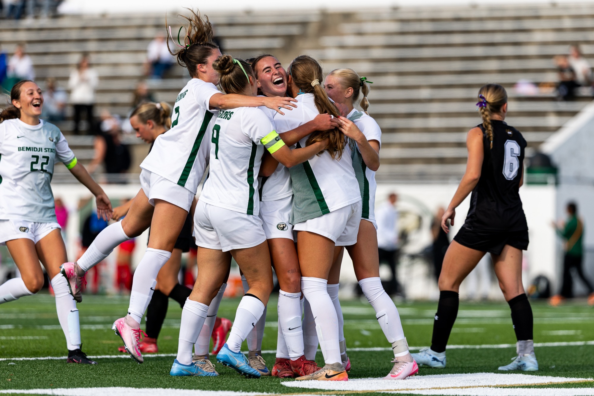 Bemidji State women’s soccer players celebrate together on the field after a goal, while opposing players and stadium seating are visible in the background.