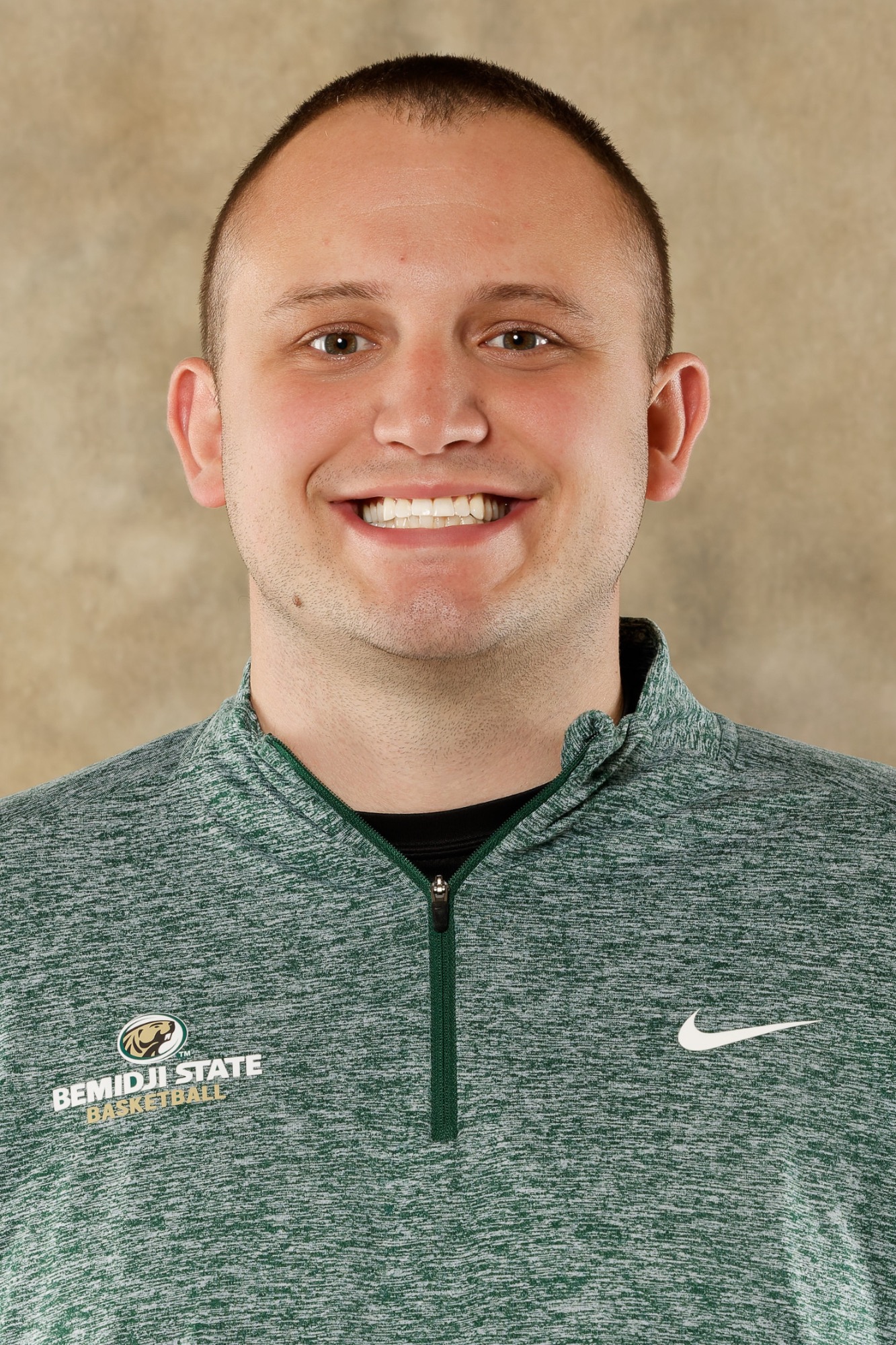 Assistant Coach Bodie Hadenfeldt poses for an official Bemidji State women's basketball staff headshot while wearing a green Bemidji State Basketball quarter-zip.