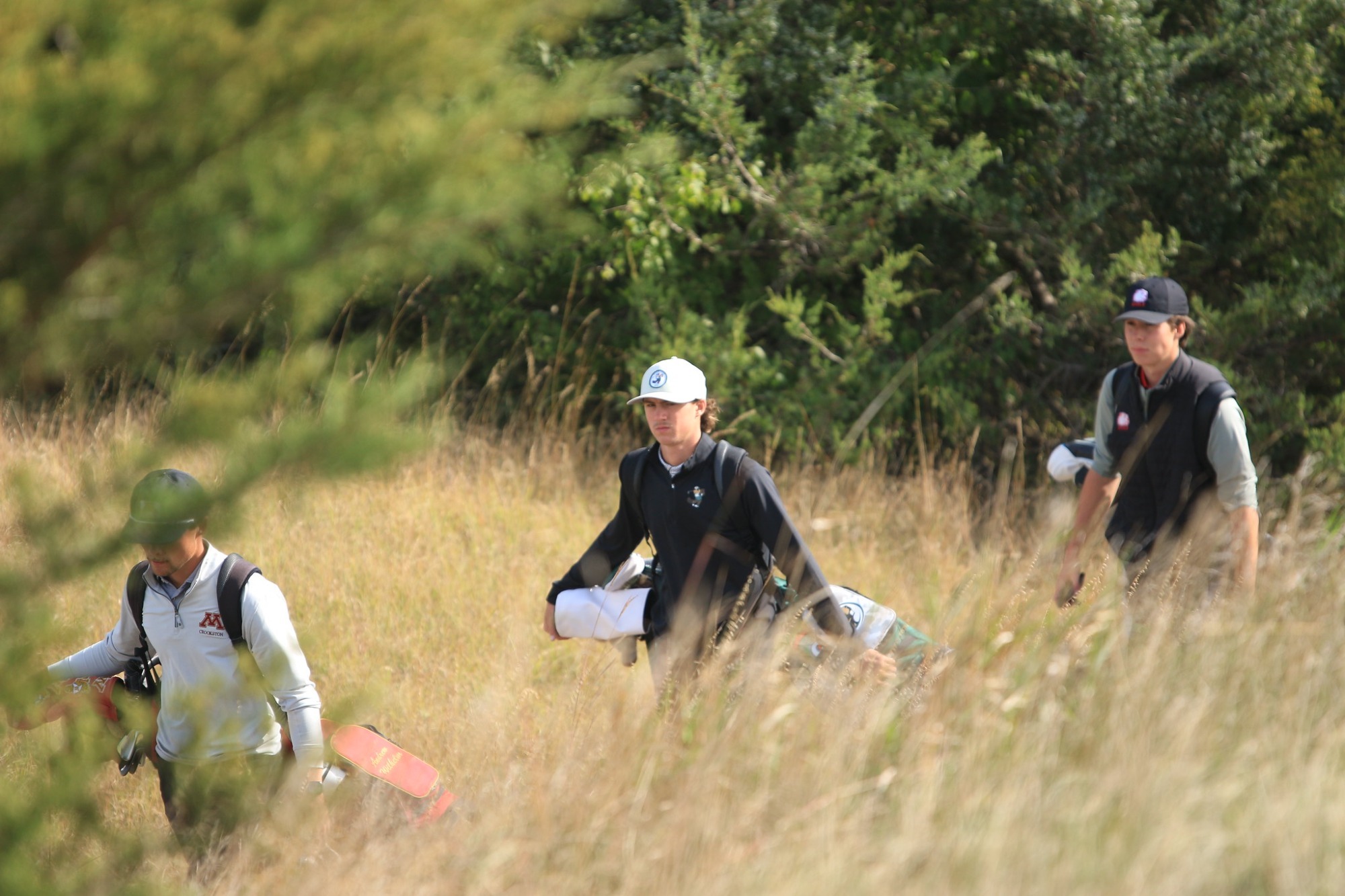 Three golfers walk through tall grass carrying their bags during a tournament round on a wooded golf course.