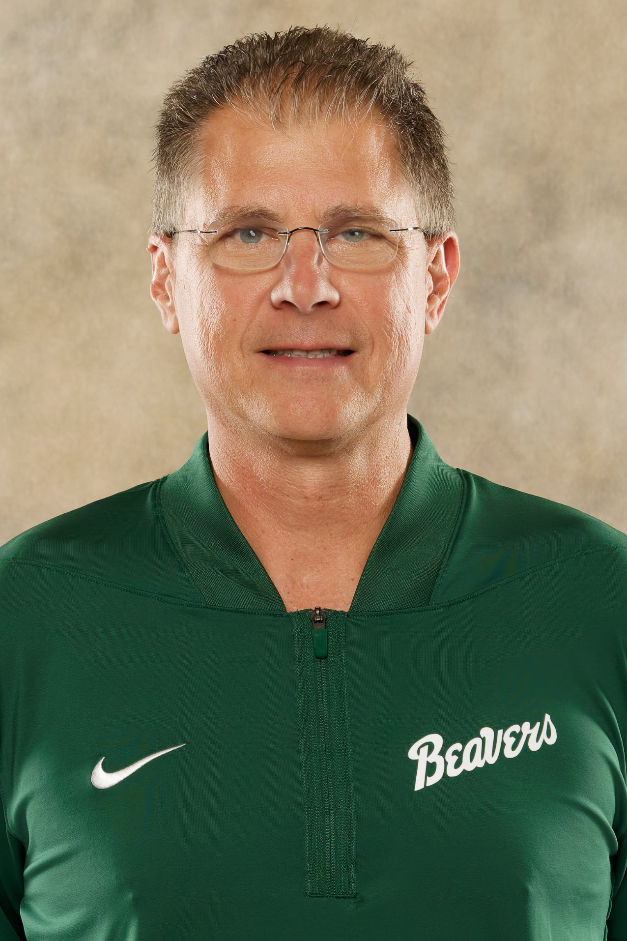 Head-and-shoulders portrait of a Bemidji State men's basketball coach wearing a green Bemidji State quarter-zip top, facing the camera against a neutral studio background.