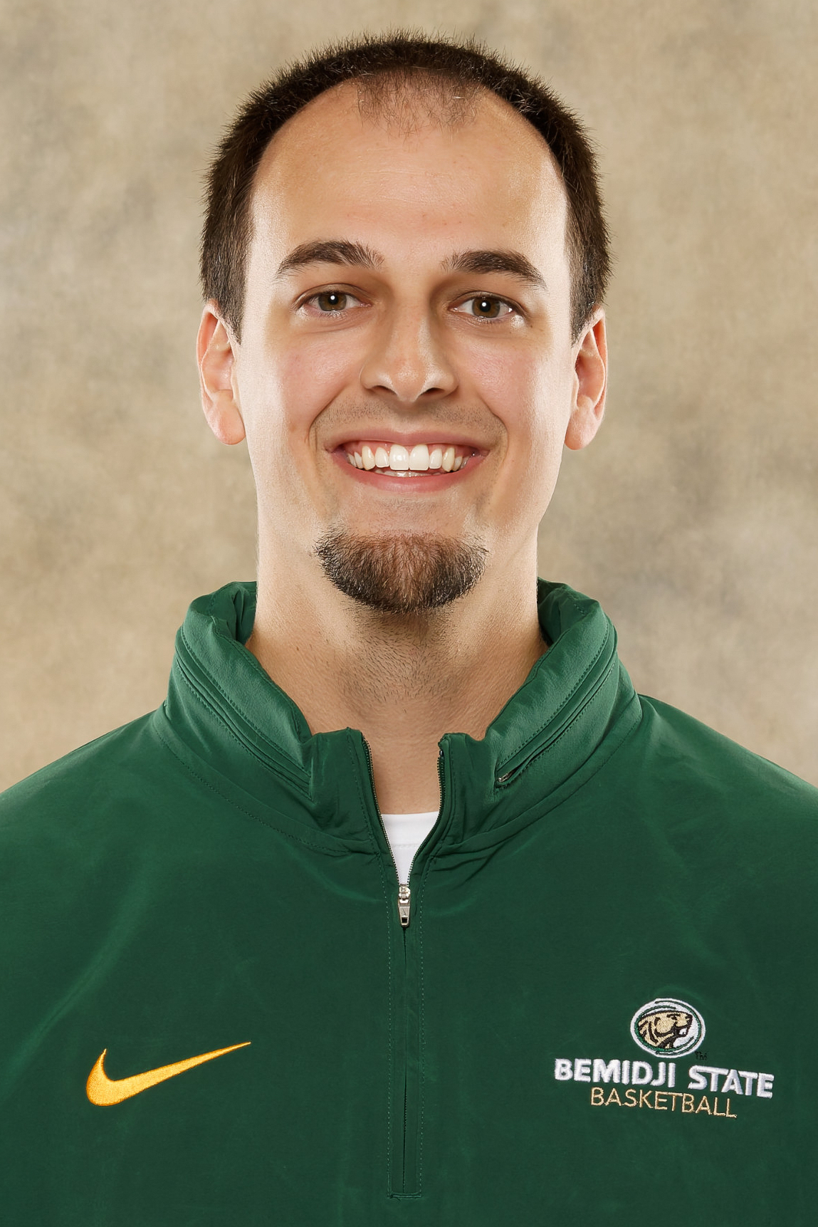 Head-and-shoulders portrait of a Bemidji State men's basketball assistant coach wearing a green Bemidji State Basketball quarter-zip jacket, facing the camera against a neutral studio background.