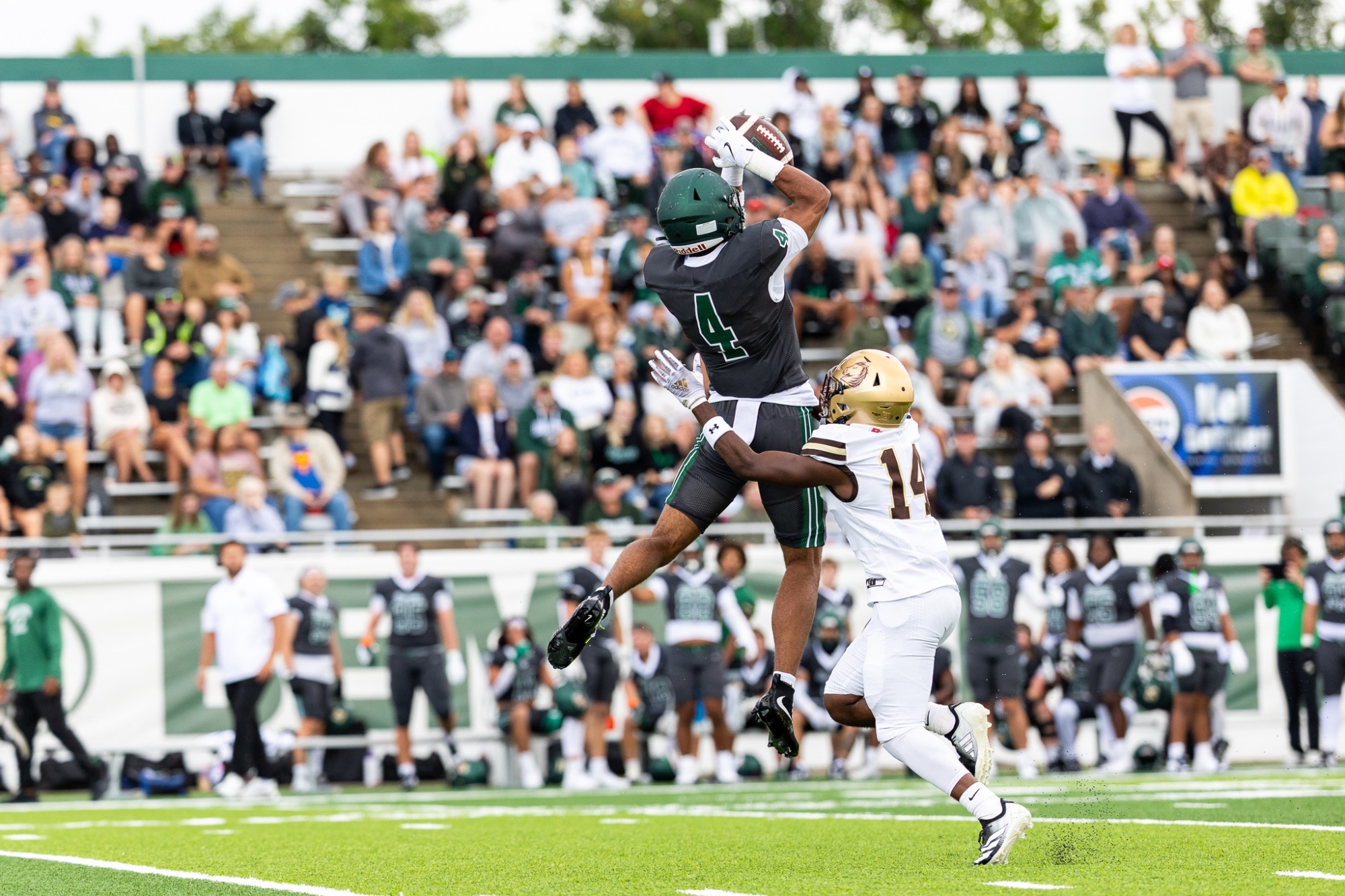 BSU's #4 Hudson Omoke leaps to catch a pass, wearing a dark grey jersey with dark green number and helmet while an SMSU defender in a white, #14 jersey with dark brown numbers and a gold helmet tackles from the ground. 