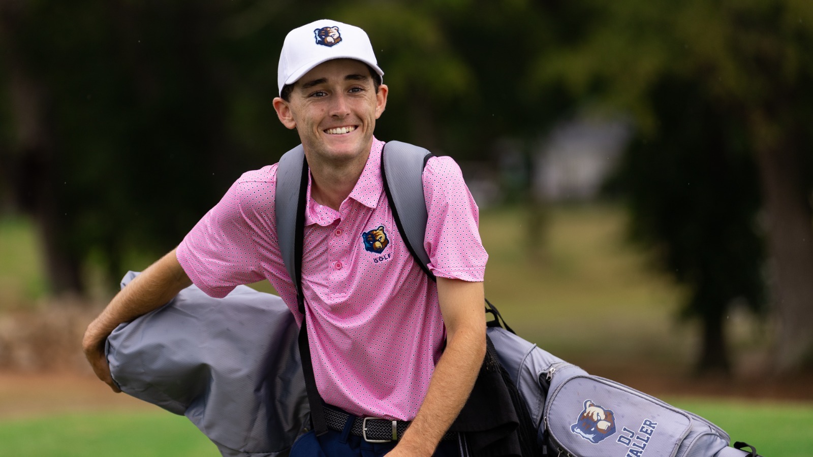 Bruins golfer in a pick polo holding his golf bag smiling at the camera