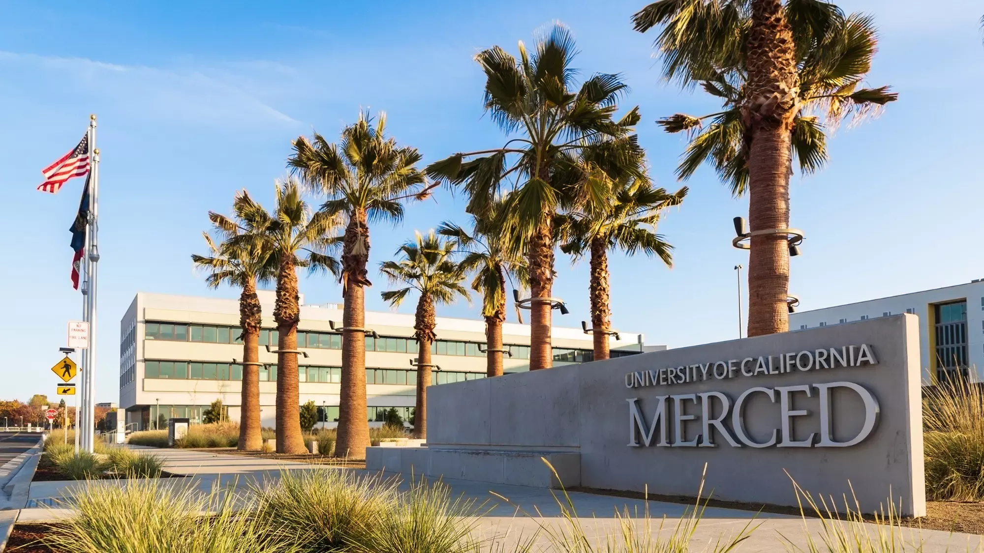 A University of California, Merced sign on University Avenue with palm trees, the American flag, and the administration building in the background.