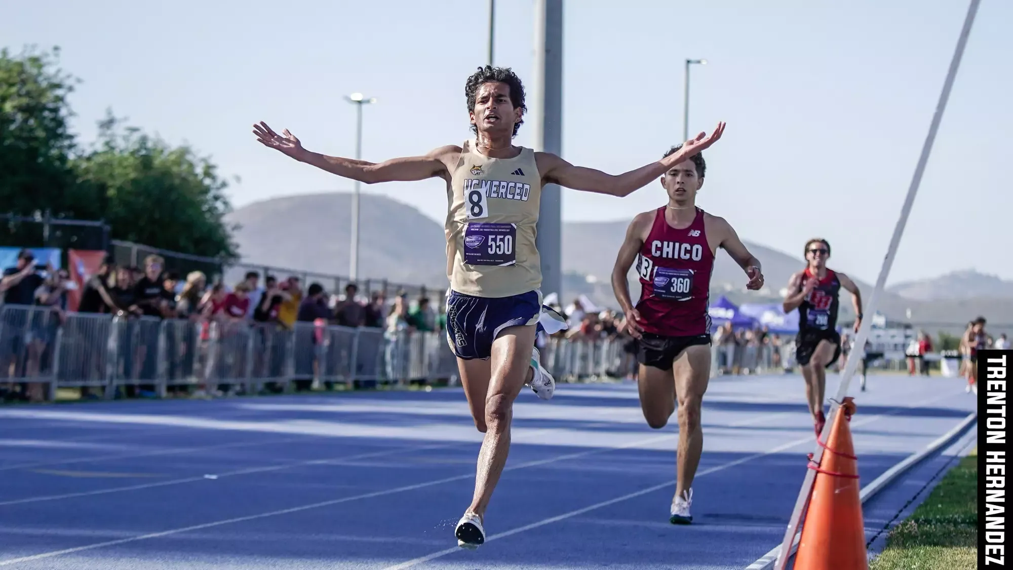 Sharvin Manjrekar crosses the finish line at the CCAA Championships a stride ahead of a Chico State challenger and a few strides ahead of a Cal State East Bay challenger. The crowd watches on at Cal State San Marcos.