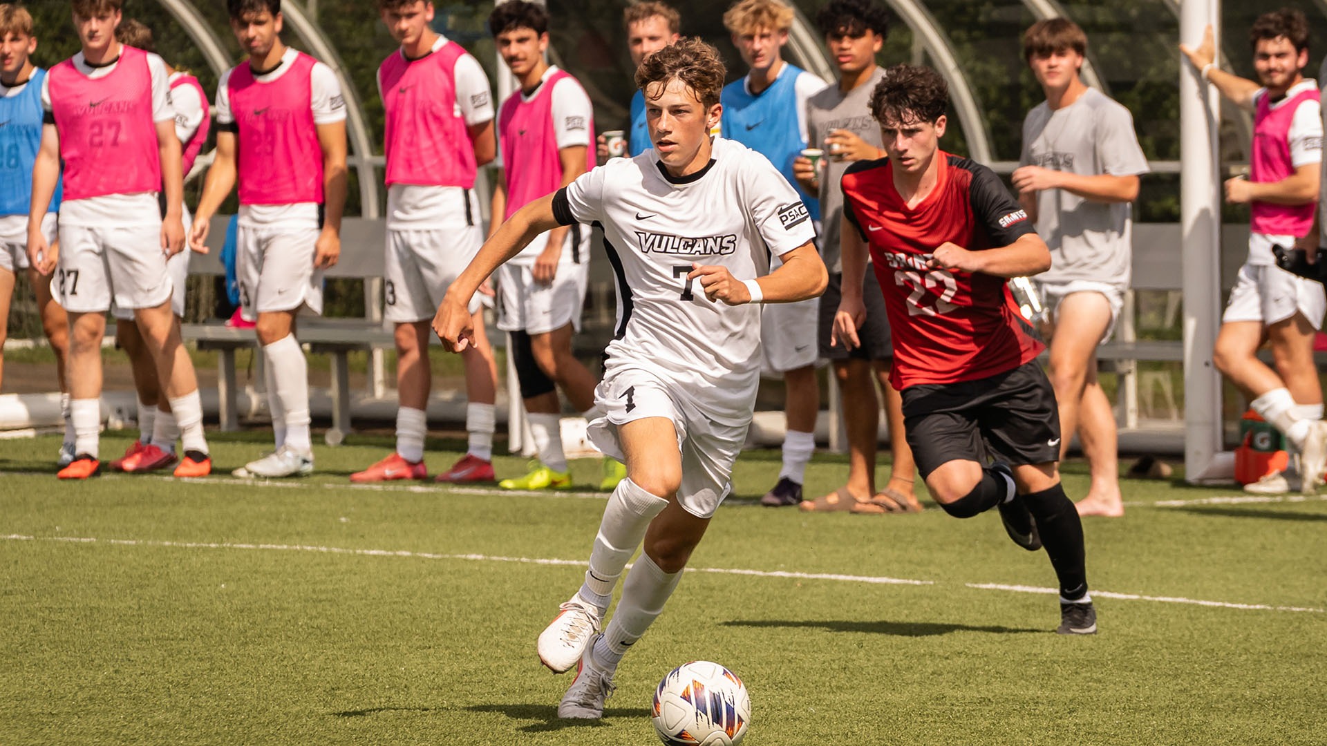 California soccer player Ryan Moessner dribbles the ball down the field in a white uniform ahead of a defender in red.