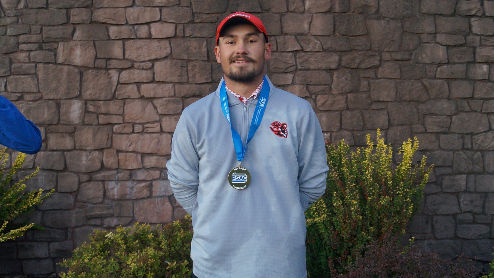 California men's golfer Nixen Erdely wearing a grey quarter-zip pulley, red Vulcans cap, and a PSAC championship medal around his neck while standing outdoors in front of a stone wall.