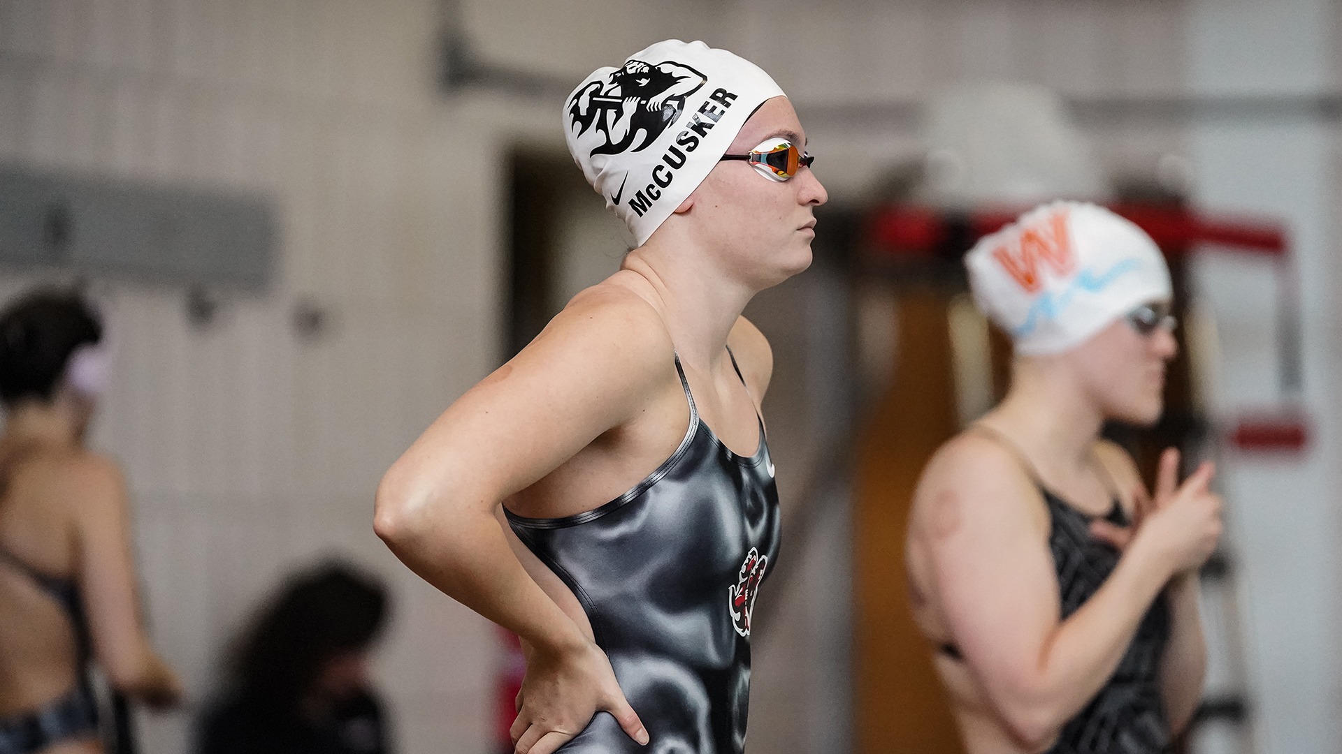 Adeline McCusker stands poolside wearing a branded swim cap and goggles, focused prior to a swimming event.