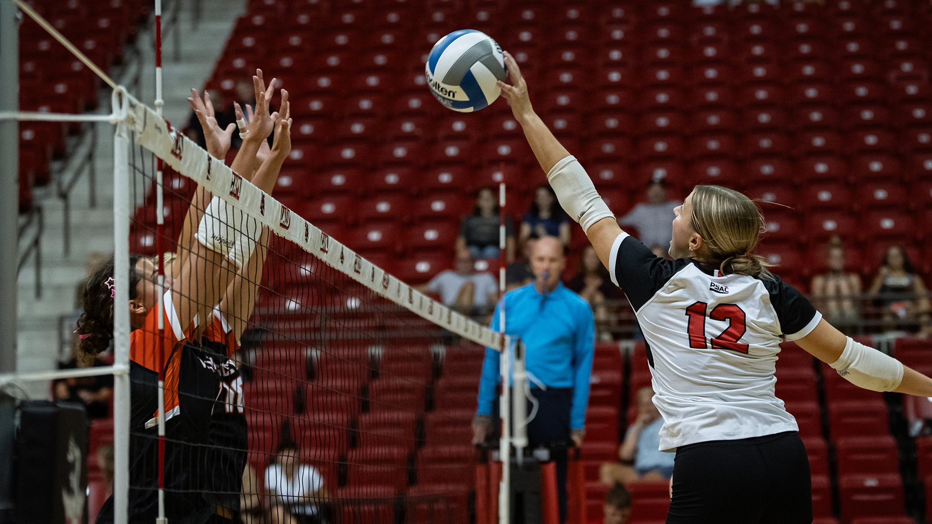 Olivia Weiseman, wearing a white and black jersey with the number 12, reaches mid-air to hit a volleyball over the net during a match.
