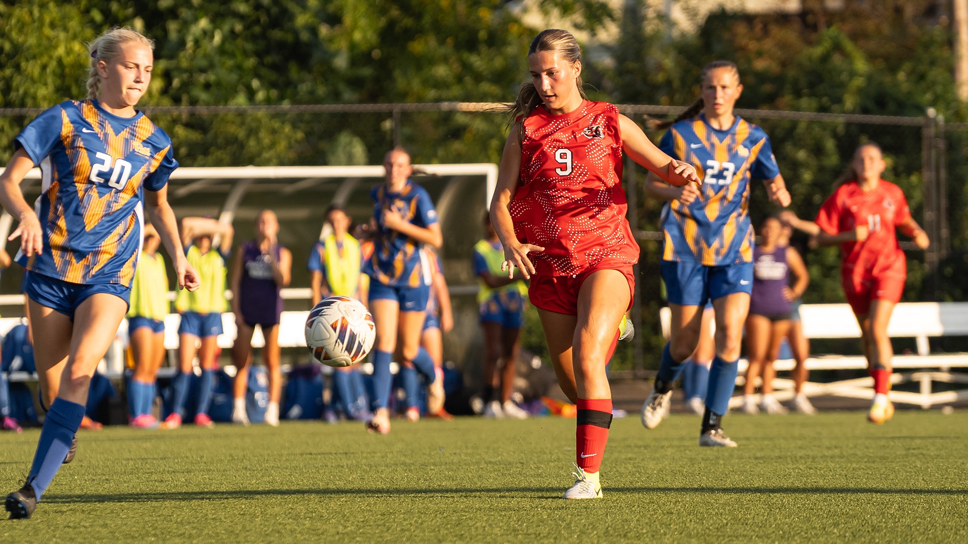 Leila Benedetti, wearing a red soccer jersey with the number 9, runs toward a soccer ball during a match on an outdoor field.