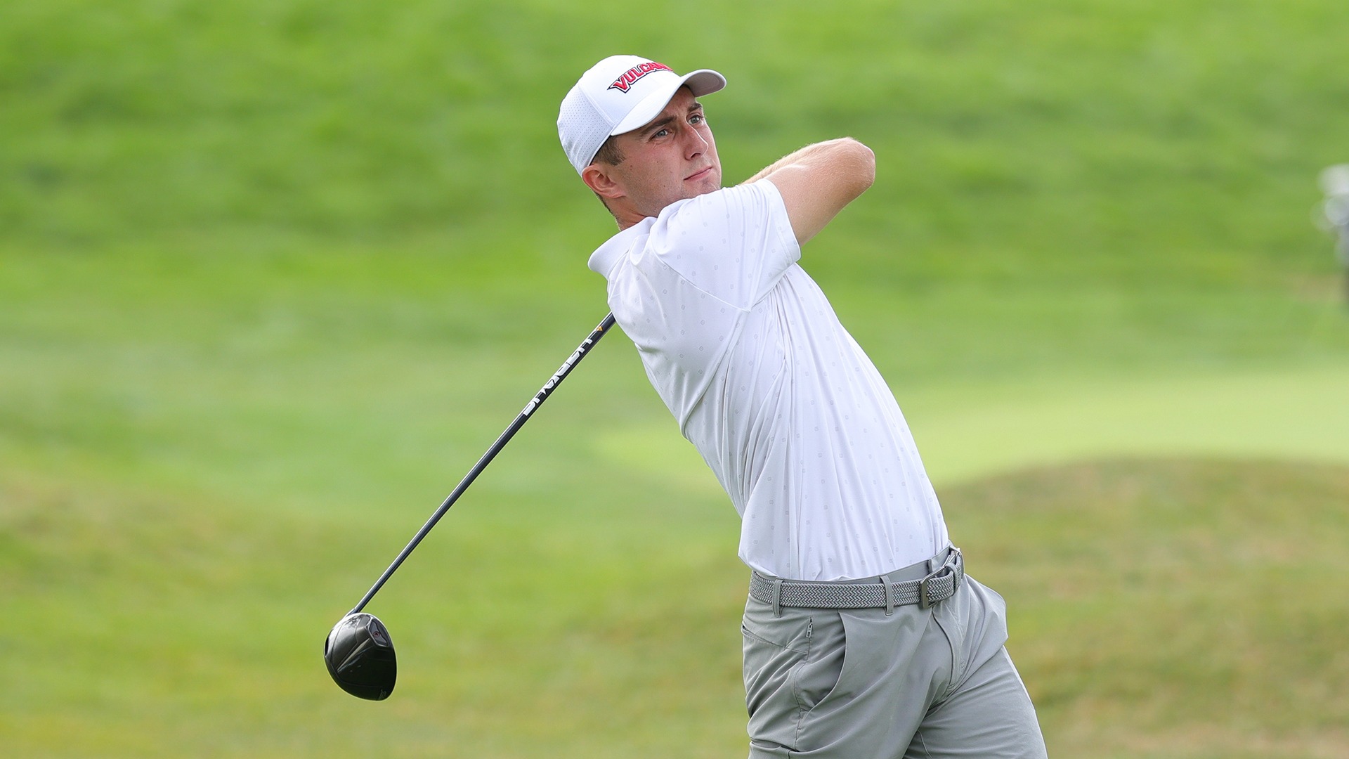 Golfer Chaz Ekoniak watches his shot following through during a swing on a green golf course, wearing a white cap and white polo shirt.