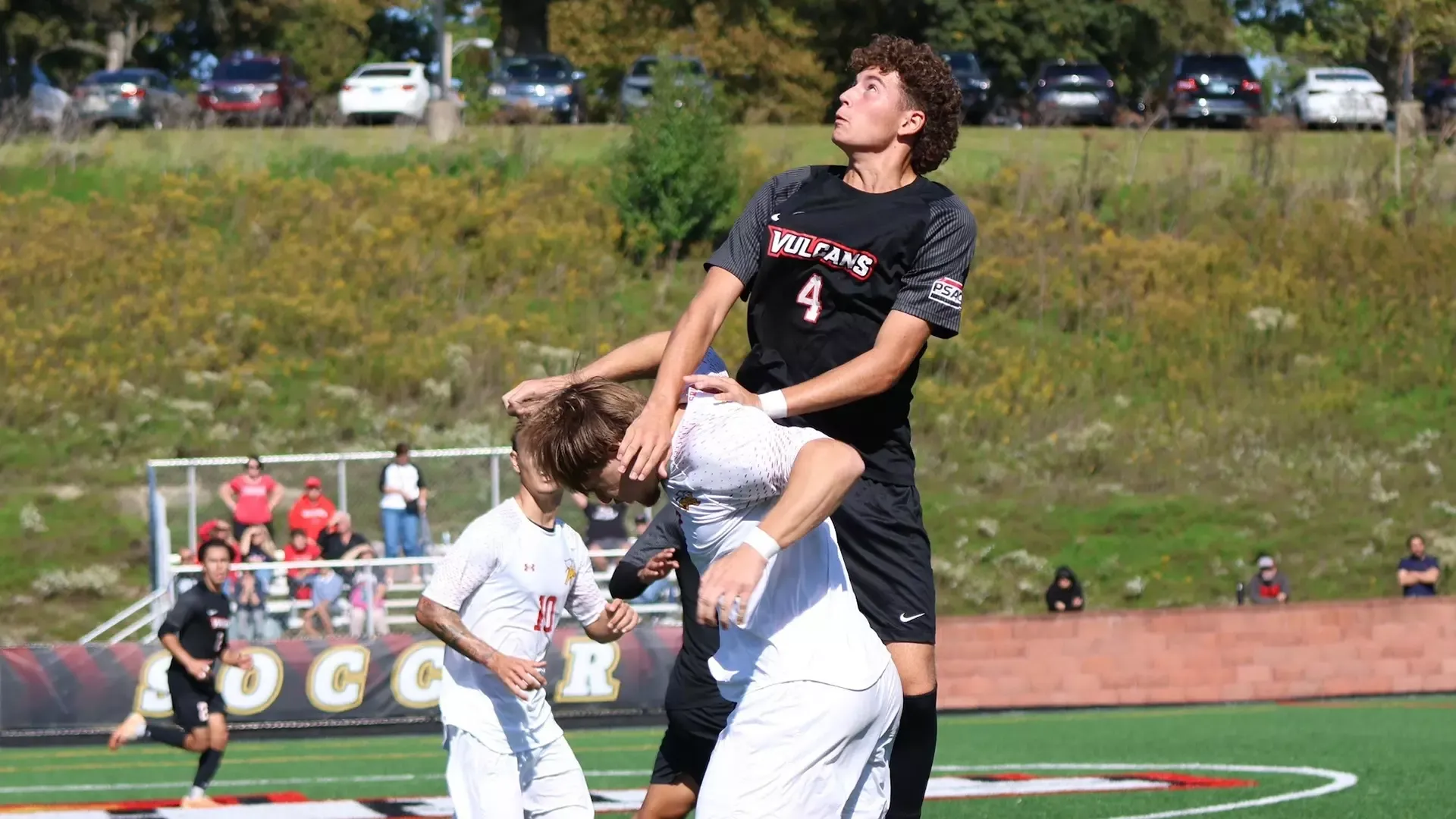 Soccer player Brayden Alvarez wearing number 4 leaps high in the air to track the ball during a daytime match on a green field.