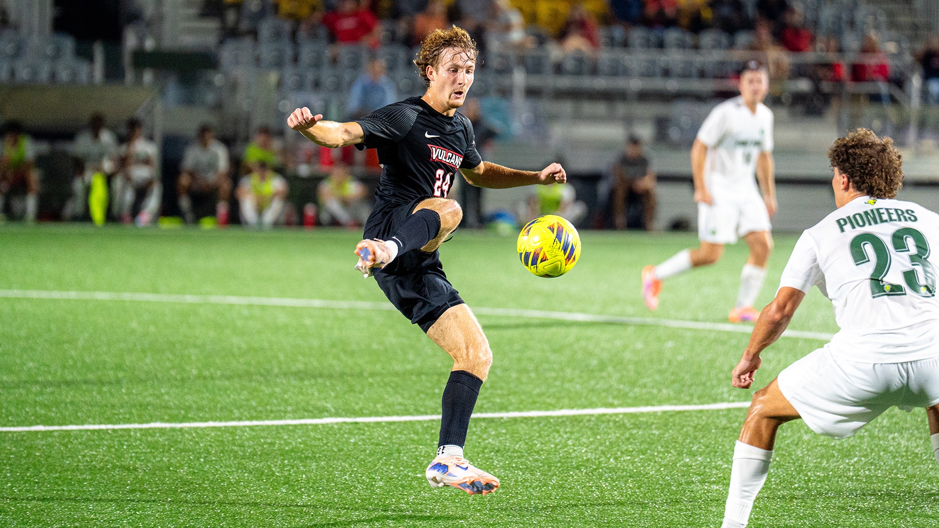 Soccer player Cole Radcliffe kicks a yellow ball mid-air during a night game on a turf field while an opposing player watches.