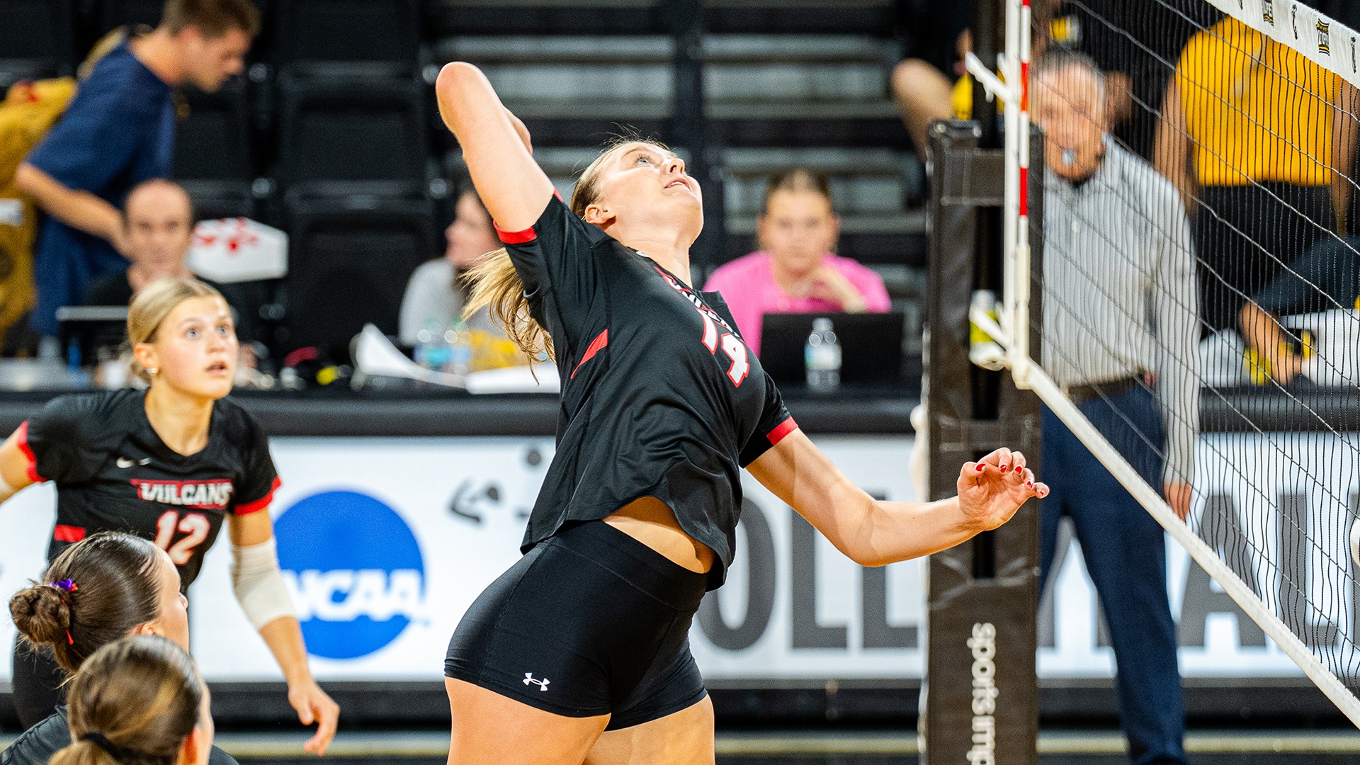 Volleyball player Abby Tucker leaps mid-air to spike the ball while teammates and officials watch from the court.