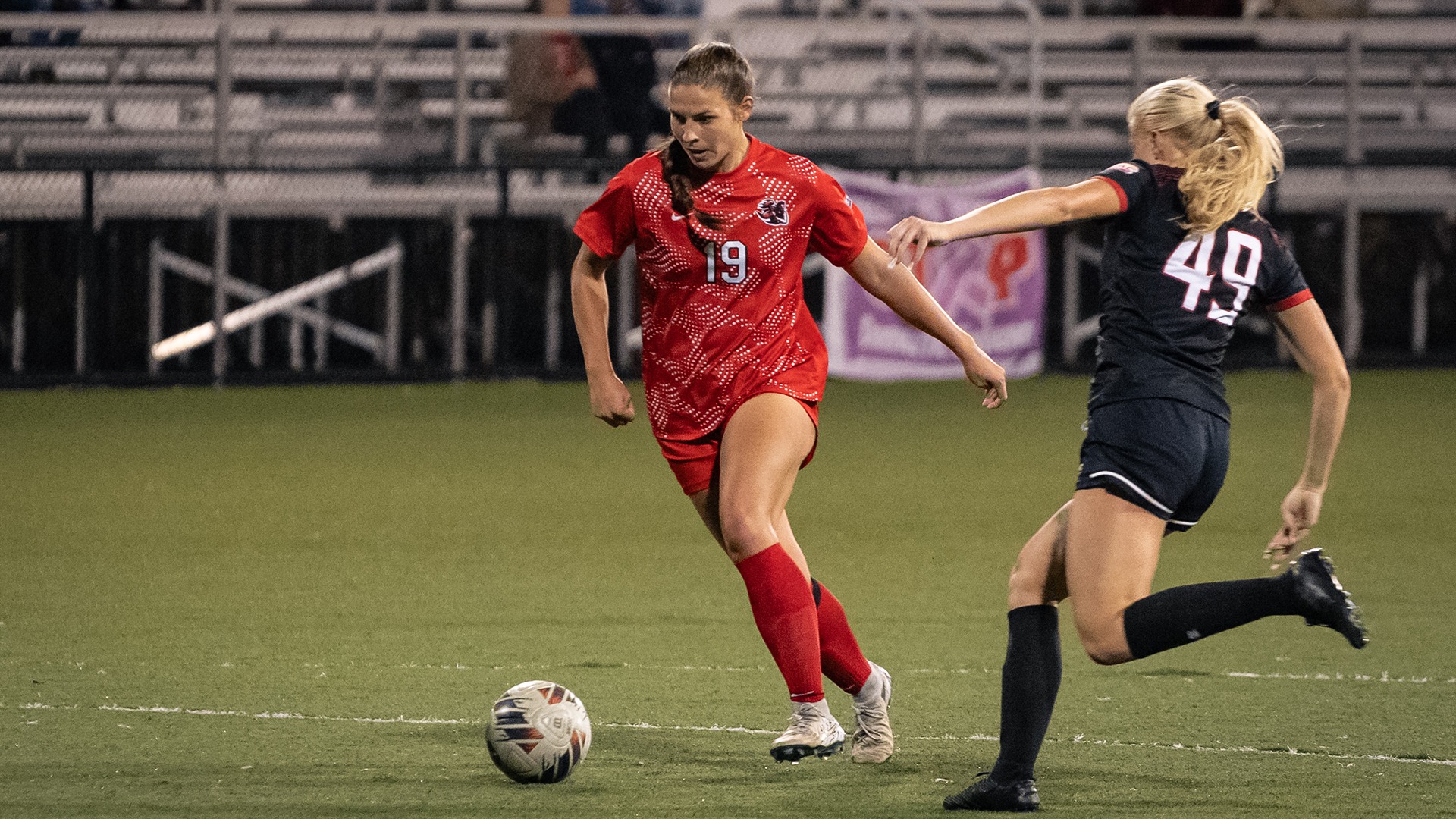 Soccer player Alexis Brown dribbles a soccer ball down the field in a red jersey with number 19 as a defender pursues her.
