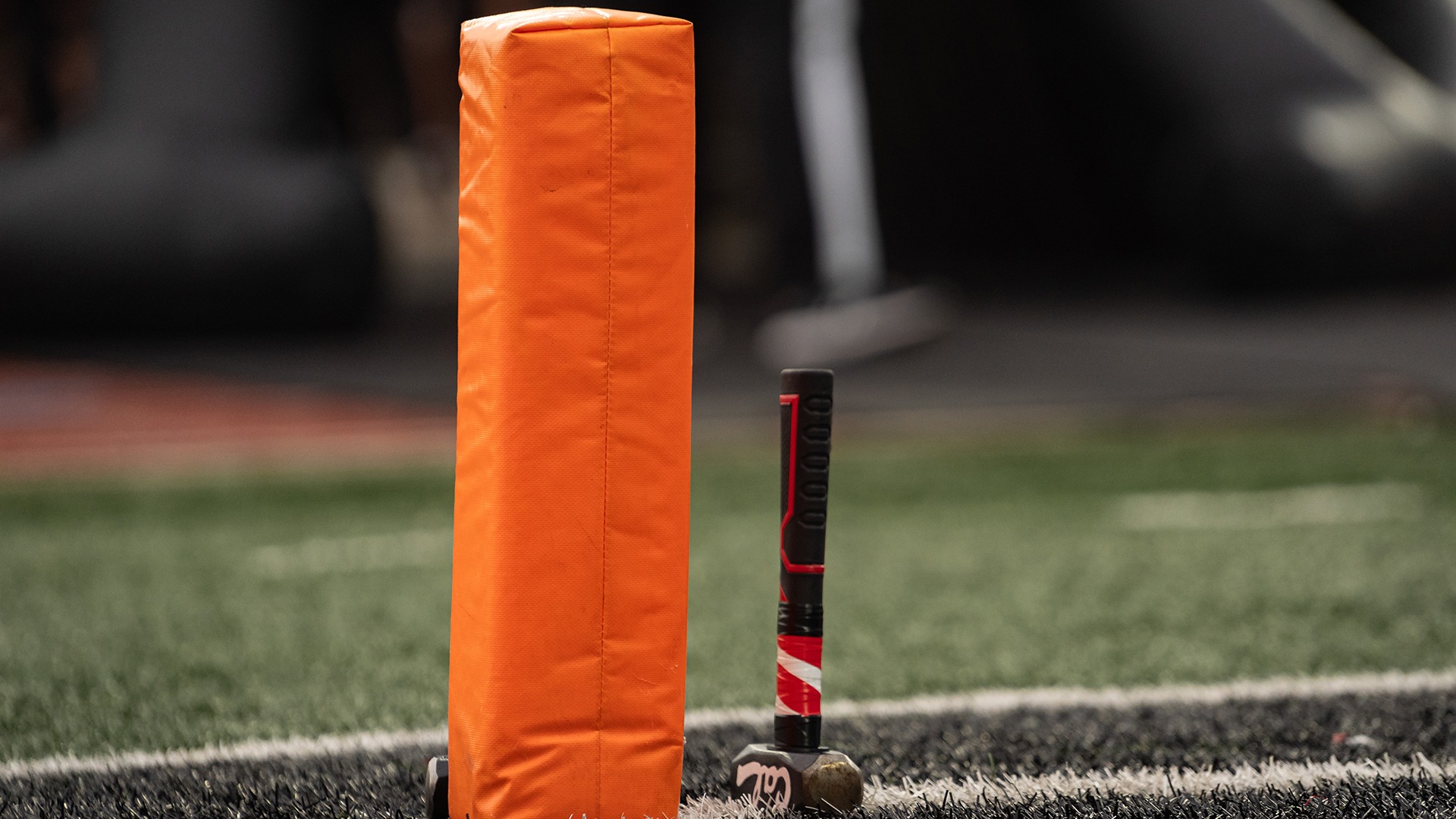 Close-up of an orange pylon and a sledgehammer with black and red grip sitting upright on the sideline turf of a football field.