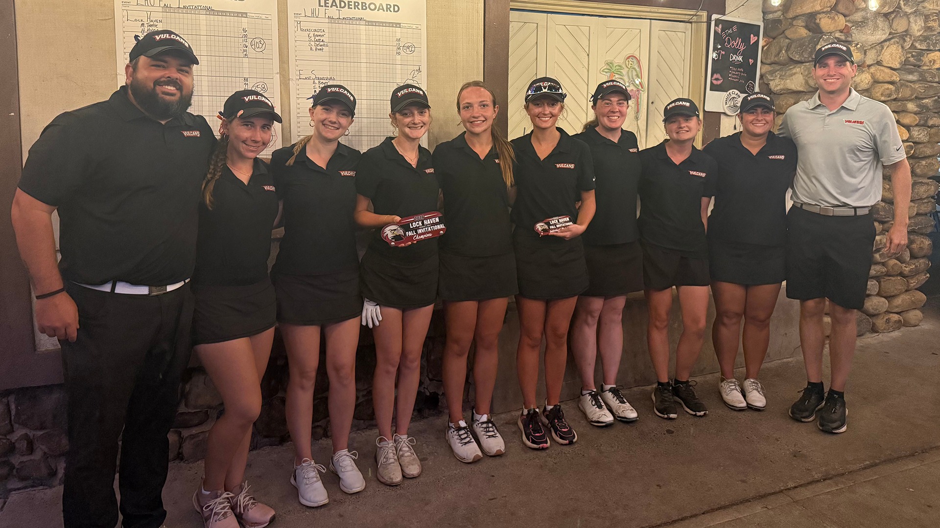 Members of the women's golf team and coaches pose together while holding awards in front of a leaderboard after winning the team title at the Lock Haven Invitational.