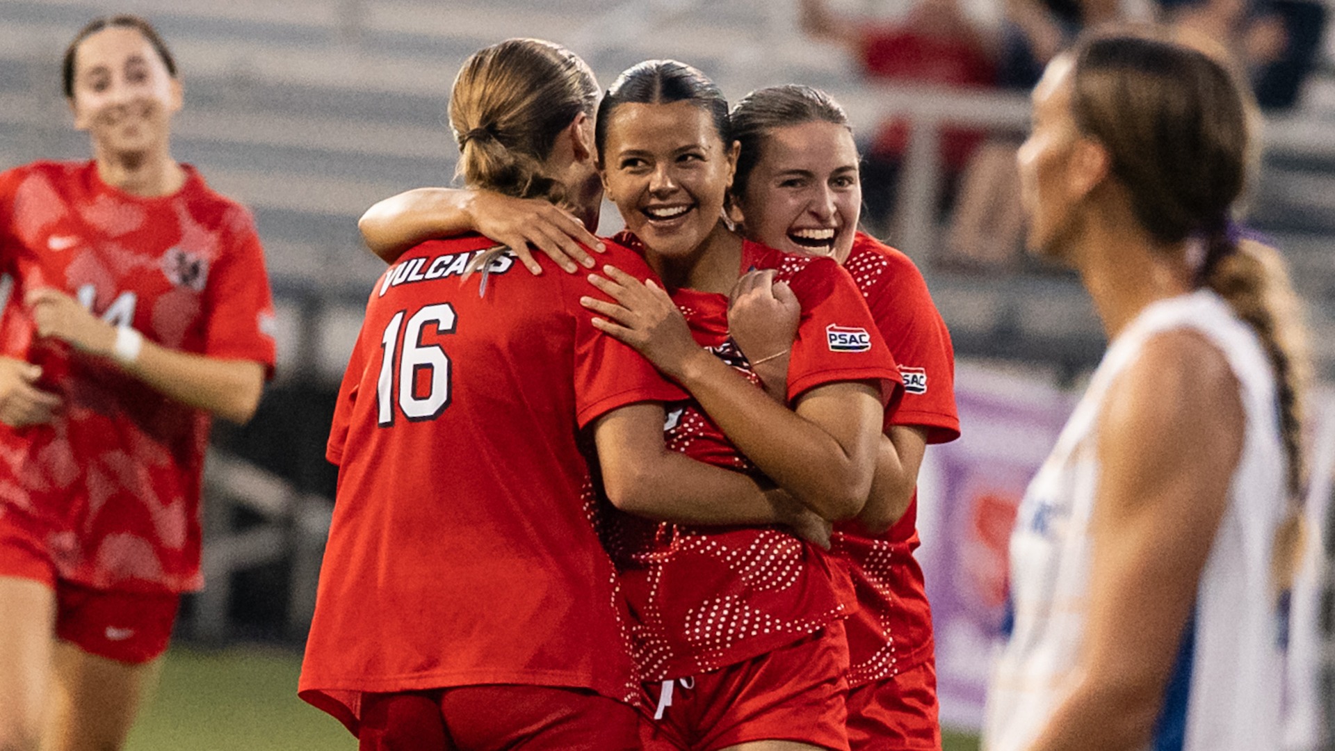 Alicia Green smiles and embraces teammates in red uniforms on the field while celebrating a goal against Ursuline.
