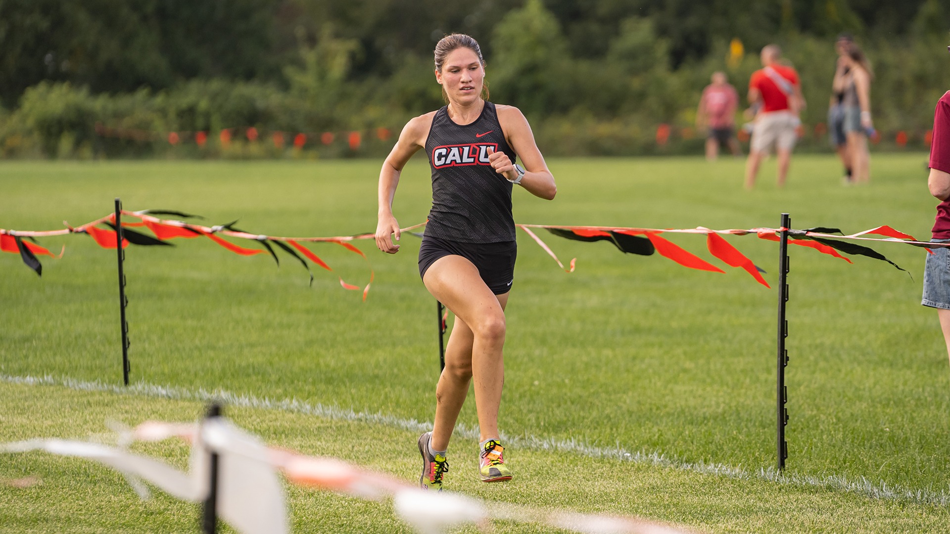 Rosalyn Perozzi running across a grassy course in a black Cal U tank top and shorts during a cross country race at Roadman Park.