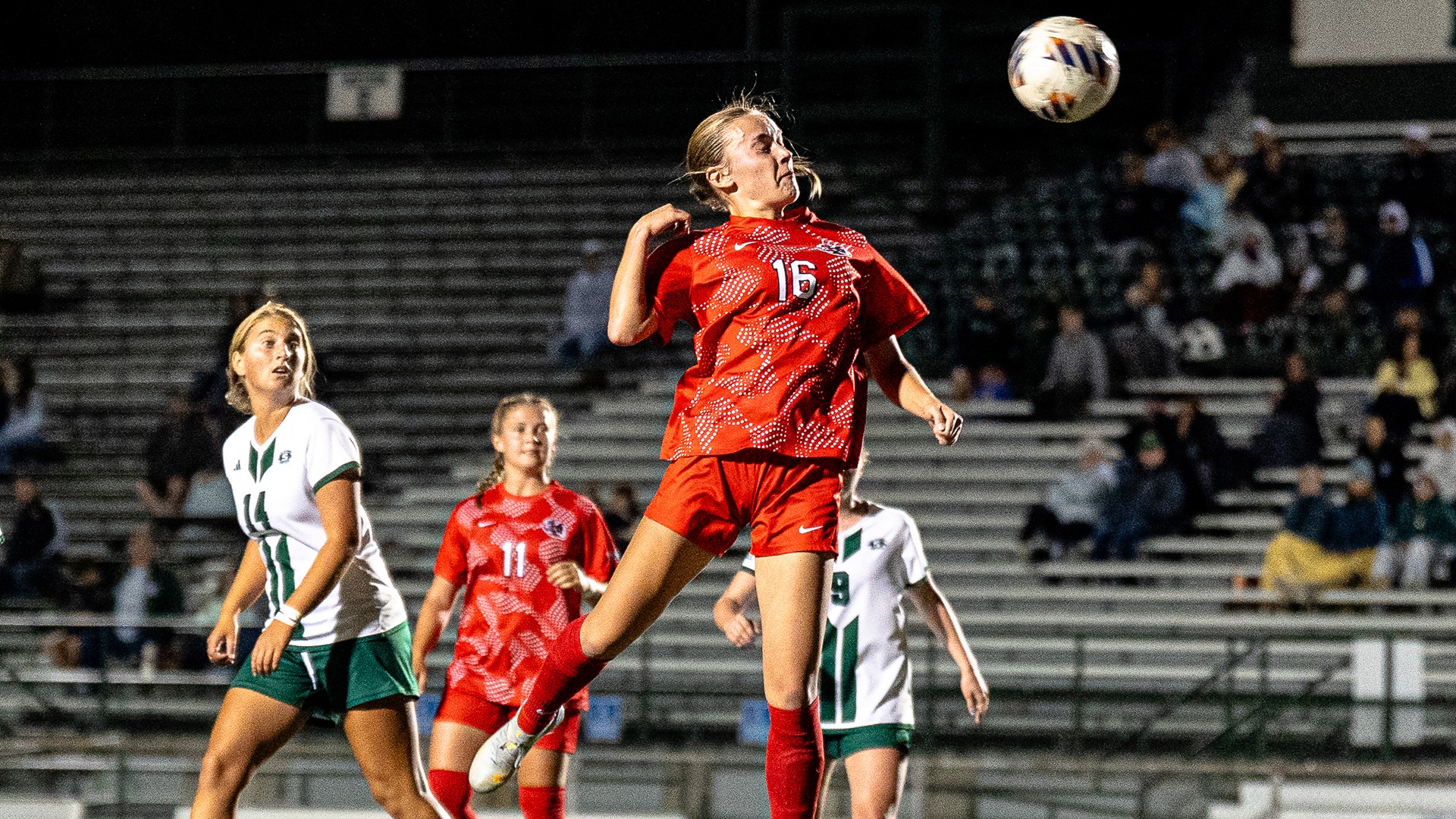Sophia Orphall leaps to head a soccer ball mid-air in red uniform during a night game against Slippery Rock.